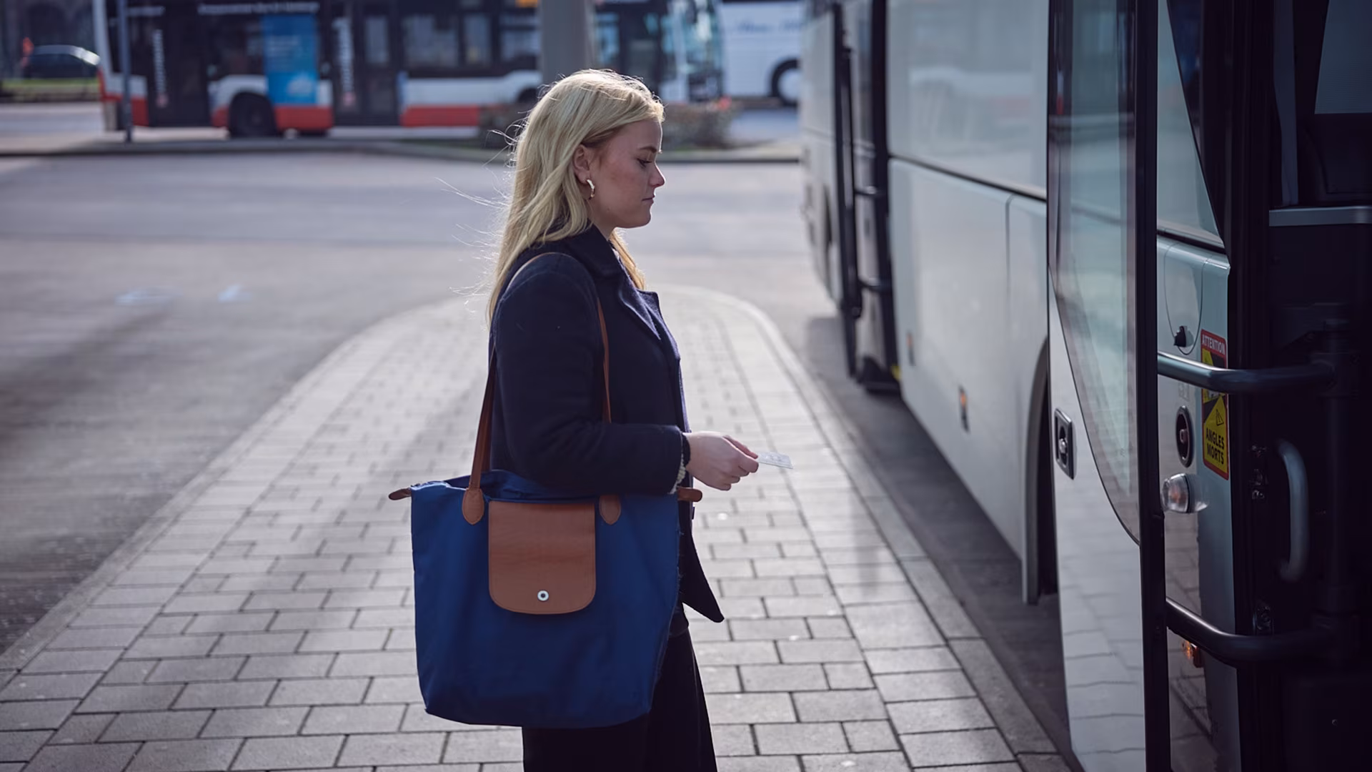Eine Frau mit blauer Tasche steigt an einer Haltestelle in einen Bus ein, während im Hintergrund ein weiterer Bus zu sehen ist.