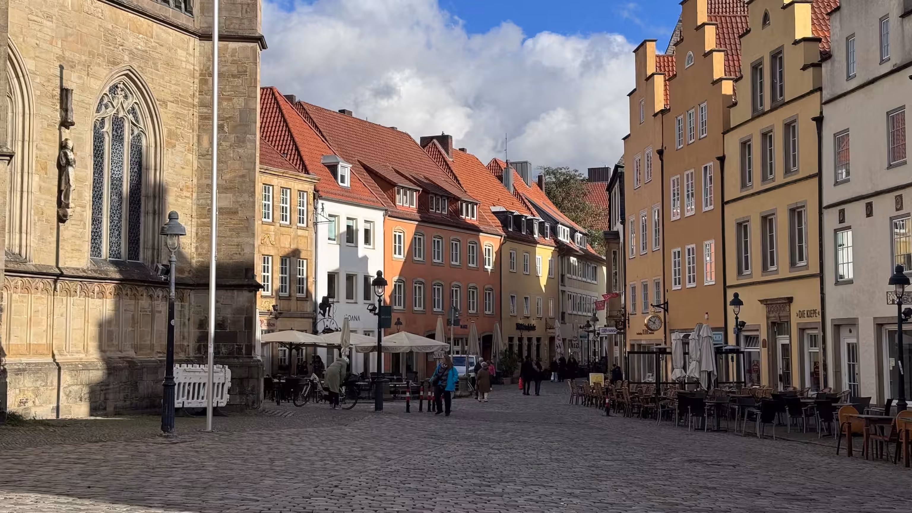 Cobbled square in Osnabrück's old town with a Gothic church façade, colourful gabled houses and outdoor dining under a partly cloudy sky.