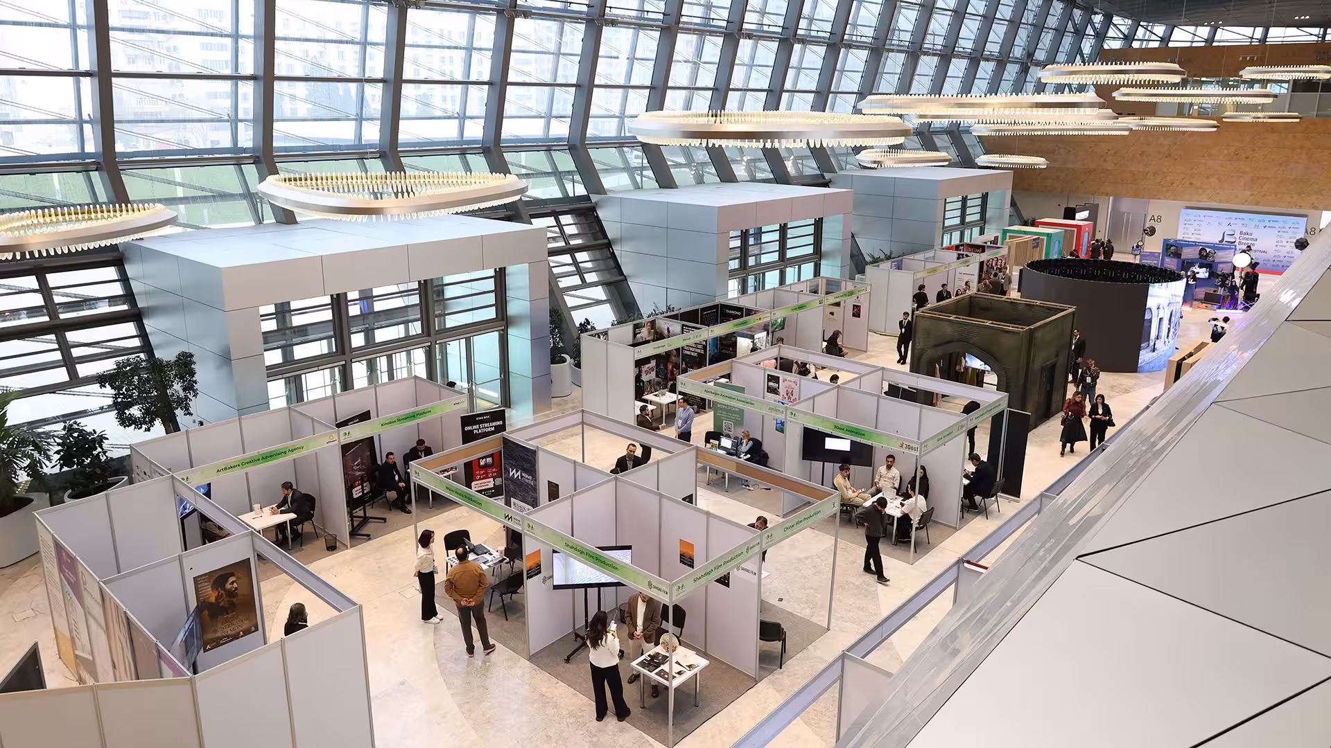 Blick von oben in eine Messehalle mit Glasfassade und weißen Messeständen, an denen Besucher mit Ausstellern sprechen.