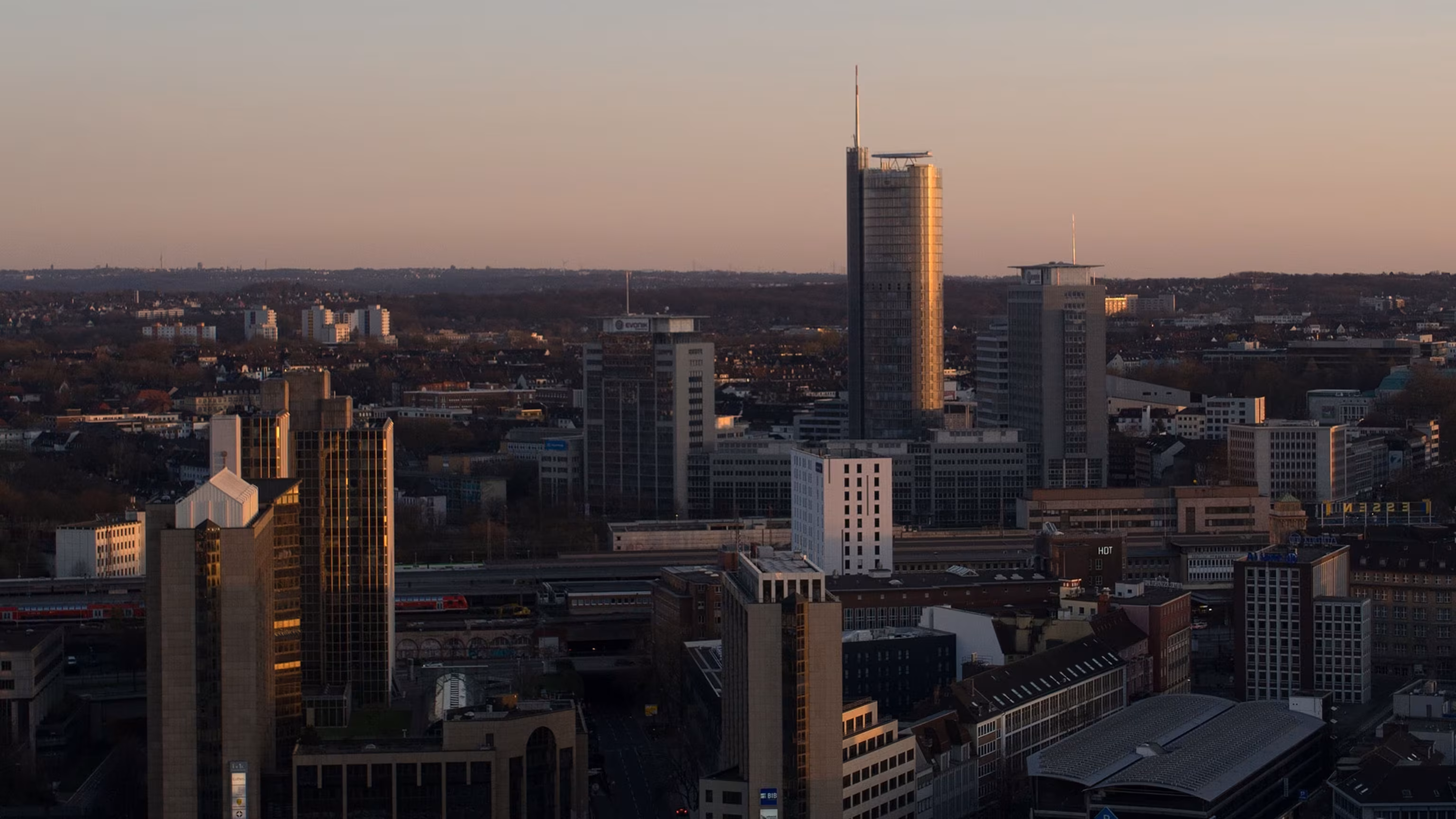 Skyline von Essen bei tief stehender Sonne: Bürohochhäuser rund um den Hauptbahnhof, in der Mitte der RWE-Tower mit Antennenmast, dahinter Wohnviertel bis zum Horizont.