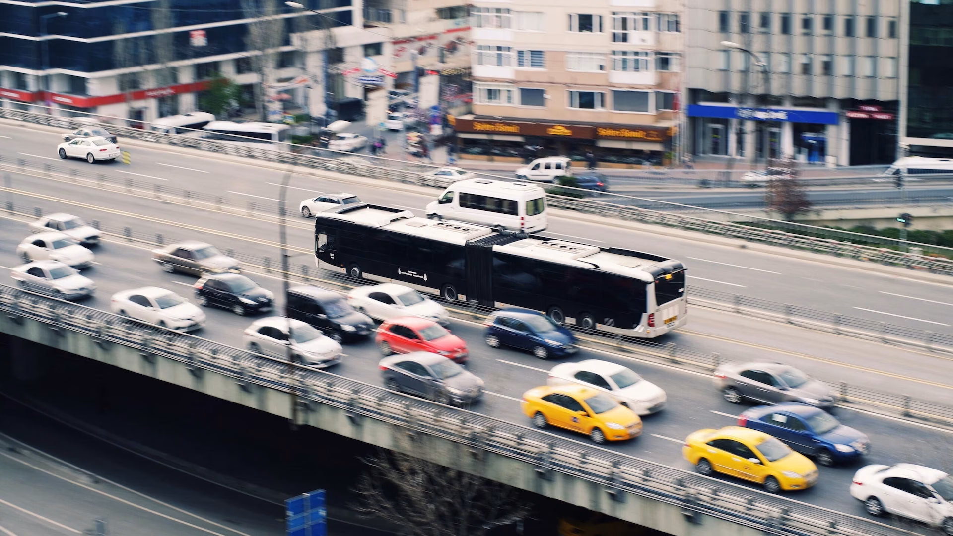 Ein langer, schwarz-weißer Gelenkbus fährt auf einer mehrspurigen Hochstraße im dichten Stadtverkehr inmitten von zahlreichen Autos und Taxis.
