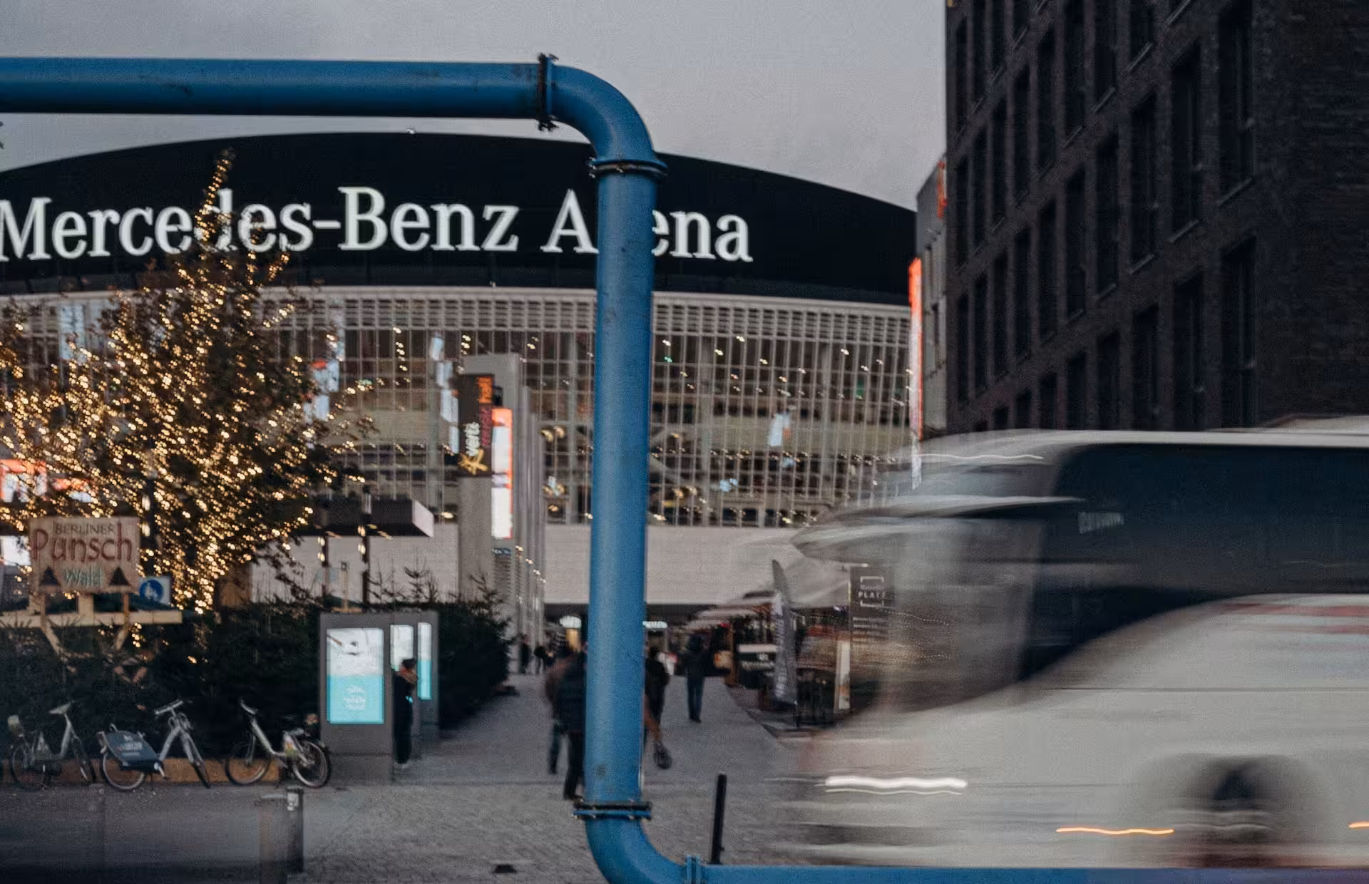 Reisebus fährt vor der Mercedes-Benz Arena in Berlin vorbei, während Besucherinnen und Besucher den beleuchteten Vorplatz in der Abenddämmerung nutzen.