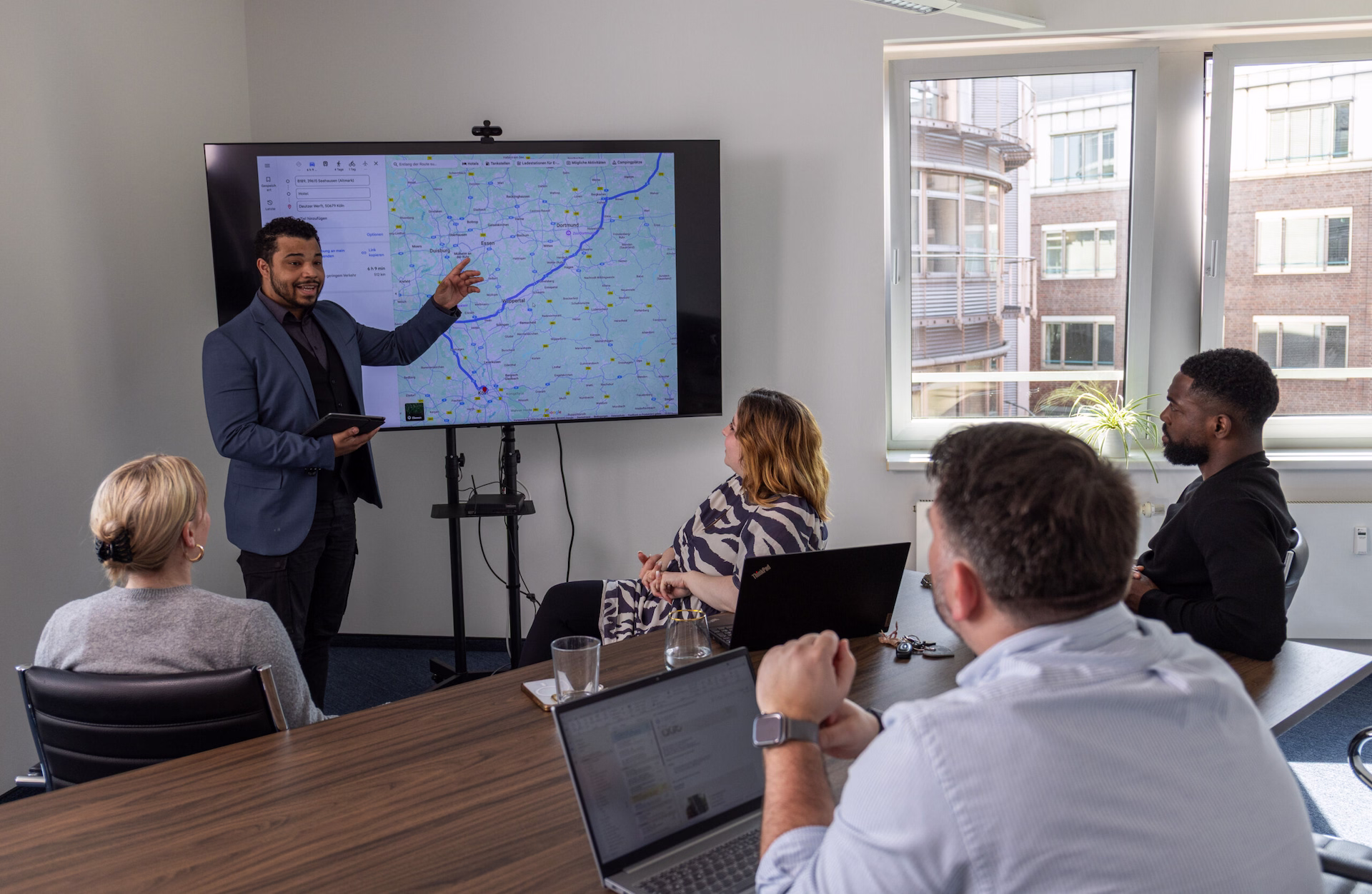 Ein Mitarbeiter präsentiert in einem Besprechungsraum eine digitale Karte auf einem großen Bildschirm. Mehrere Kolleginnen und Kollegen verfolgen die Präsentation aufmerksam und besprechen gemeinsam eine Route oder Einsatzplanung.