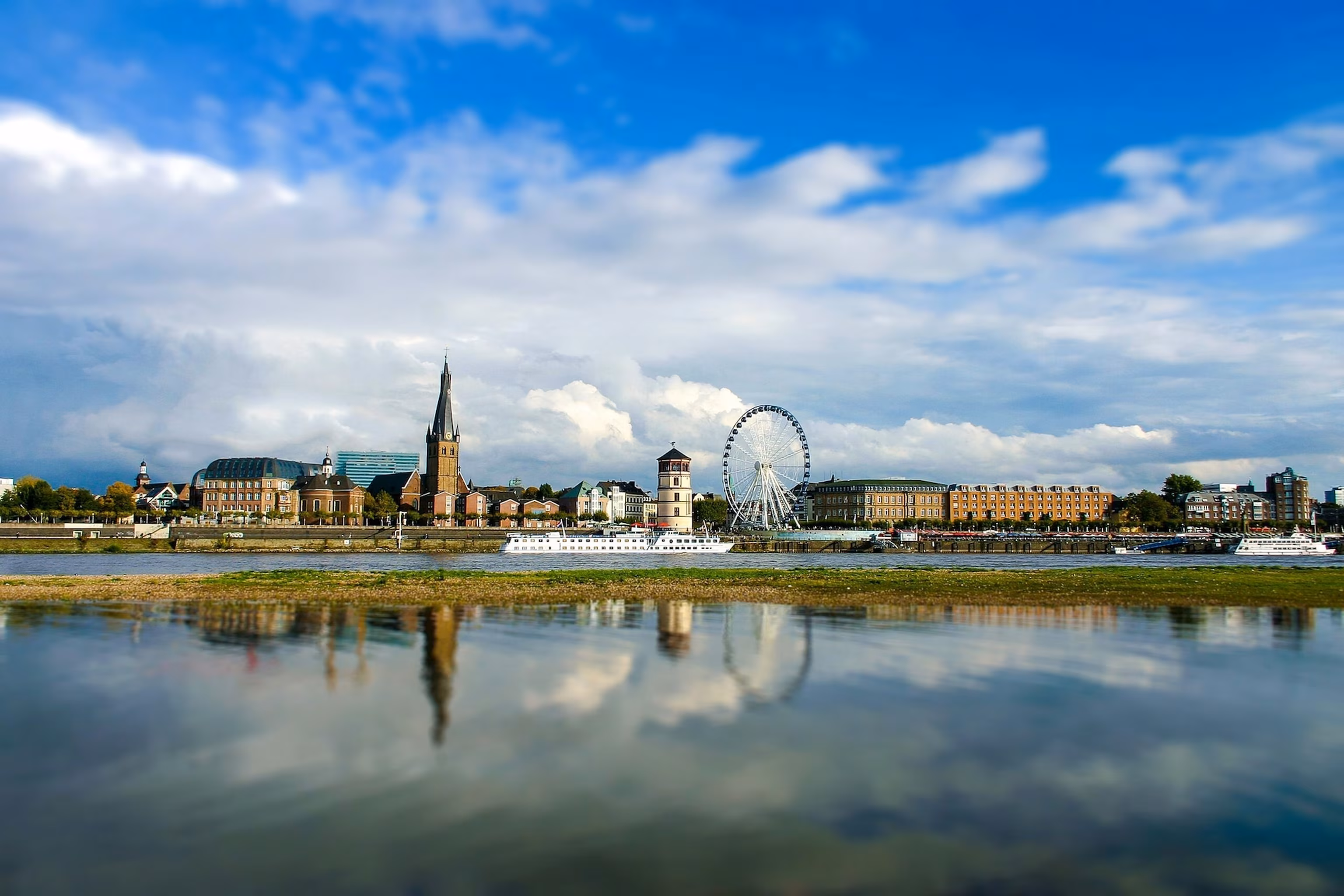 Panoramablick auf die Düsseldorfer Rheinuferpromenade mit dem Schlossturm, dem Riesenrad und historischen Gebäuden, gespiegelt im Rhein unter einem teilweise bewölkten Himmel.