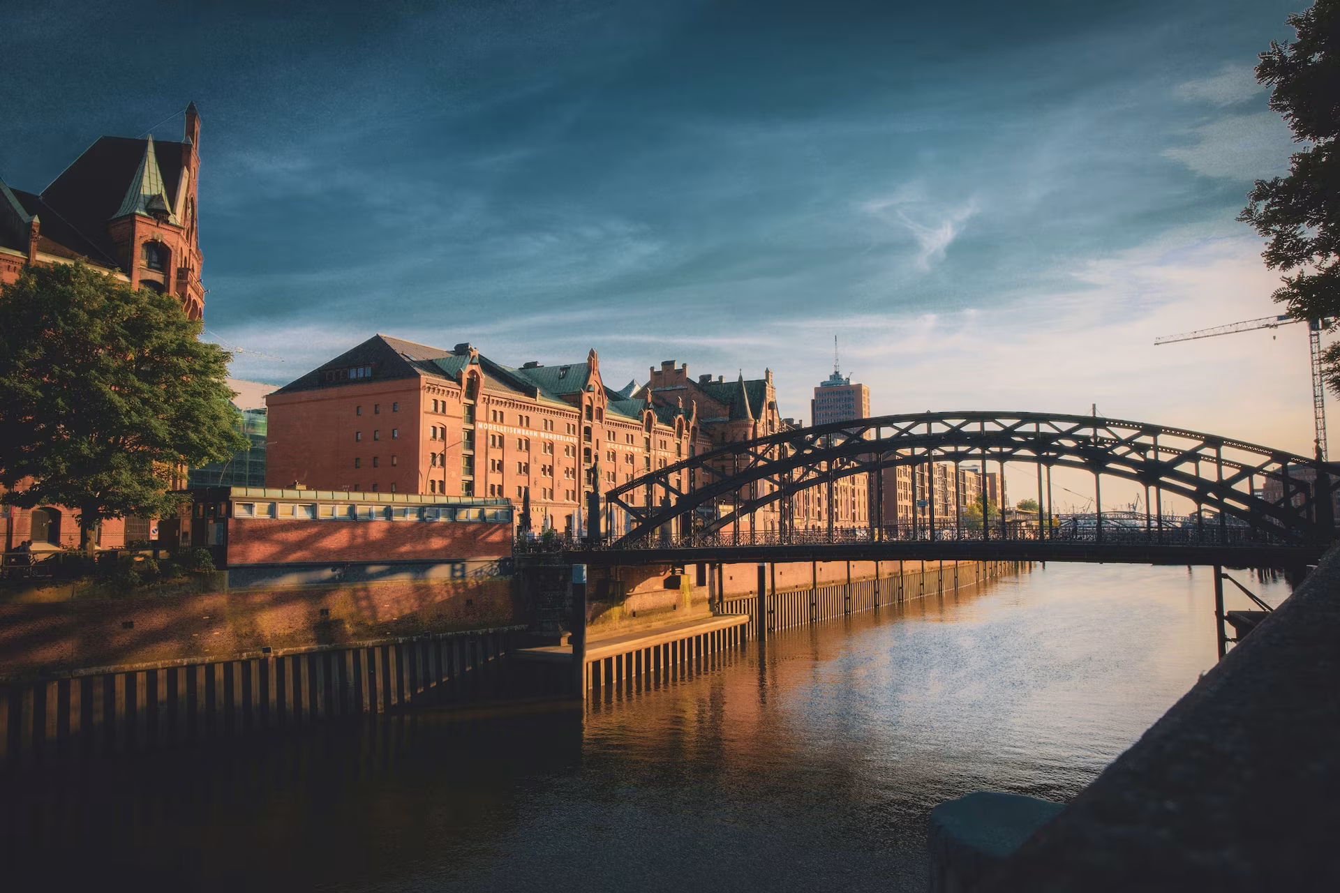 Historische Speicherstadt in Hamburg mit einer Stahlbrücke über einem Kanal, umgeben von roten Backsteingebäuden und ruhigem Wasser bei warmem Abendlicht.