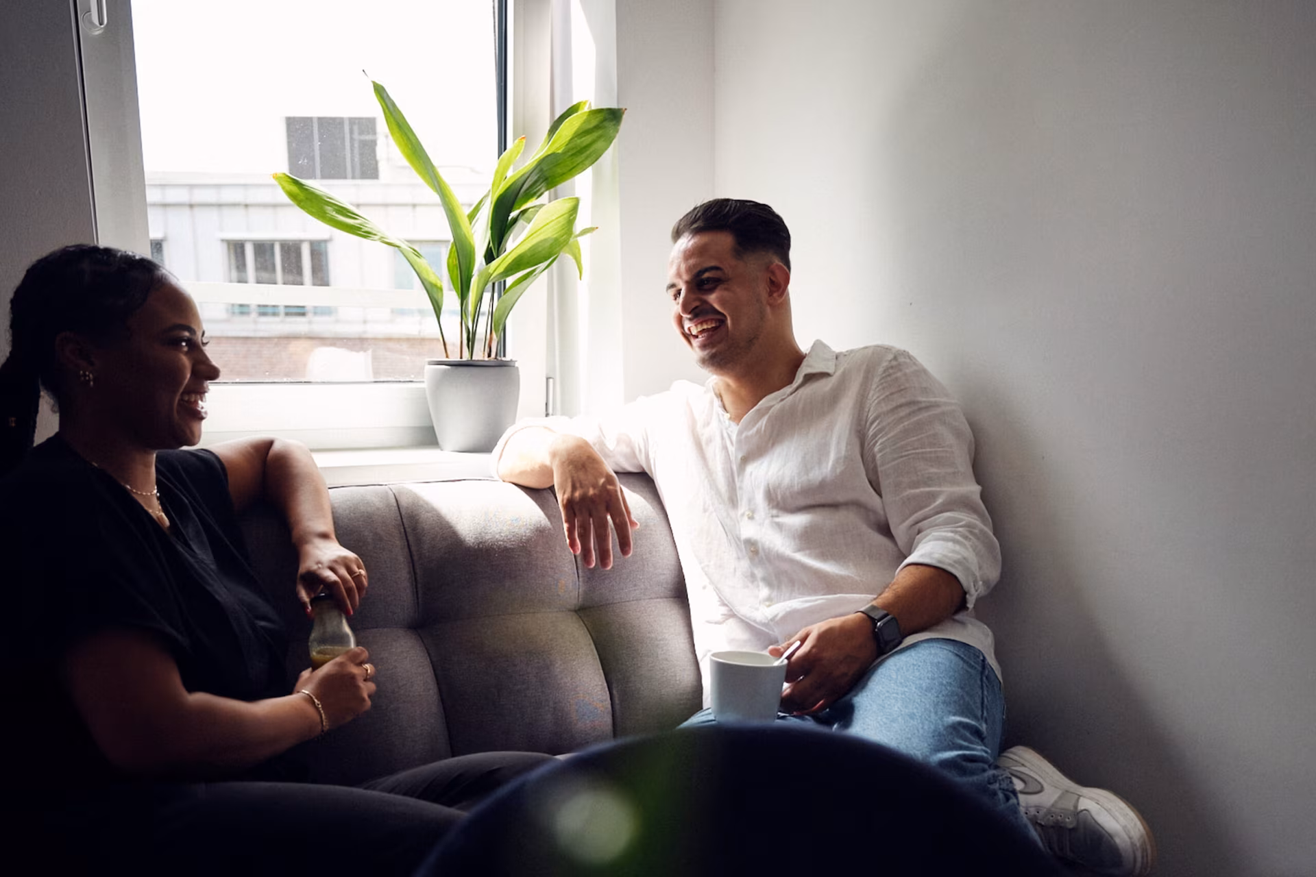 Zwei Personen sitzen auf einem Sofa nahe einem Fenster, unterhalten sich und lächeln. Auf der Fensterbank steht eine Topfpflanze.