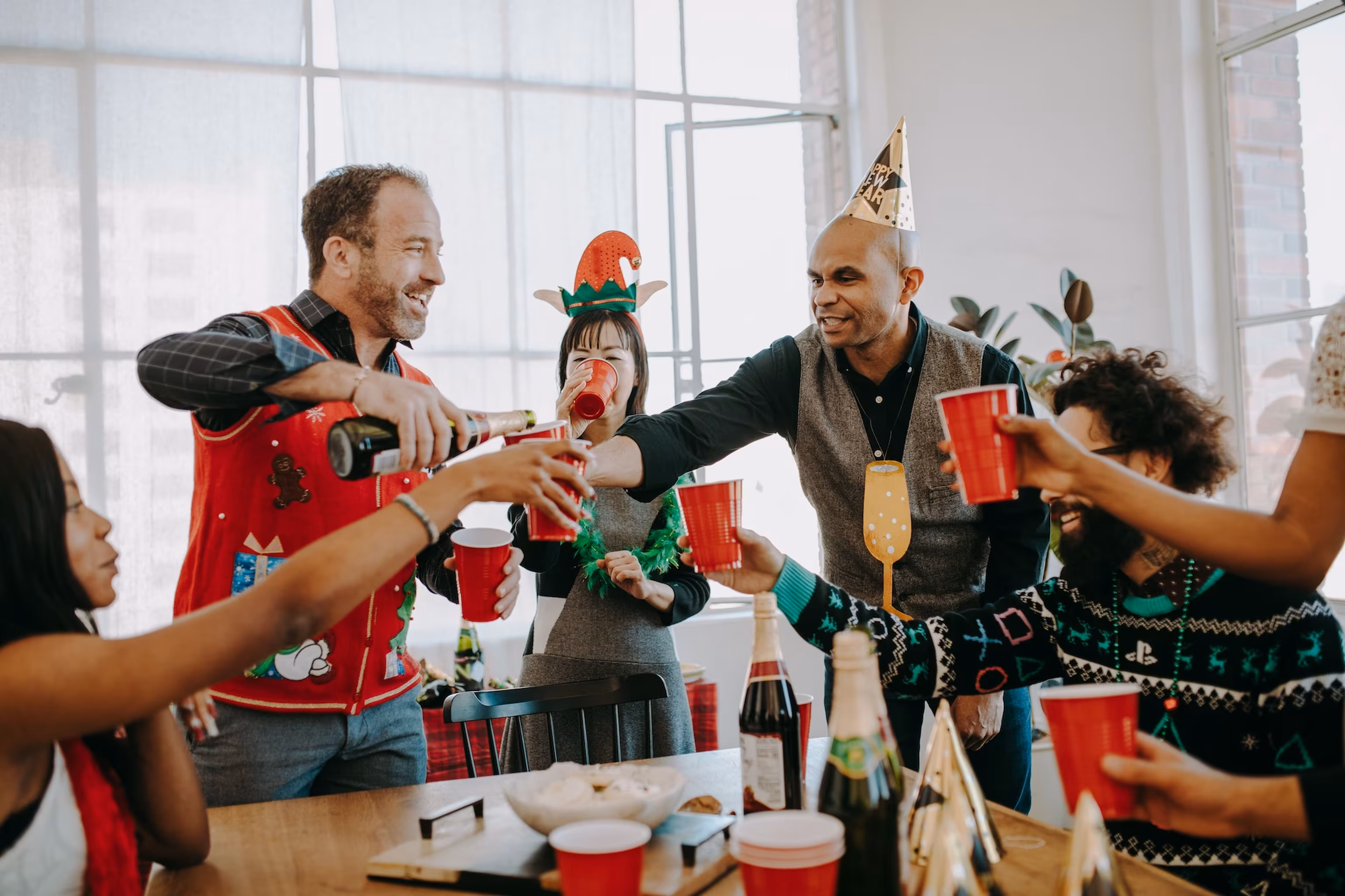 Kolleginnen und Kollegen in Weihnachtspullovern und mit Partyhüten stoßen bei einer Weihnachtsfeier mit roten Bechern an