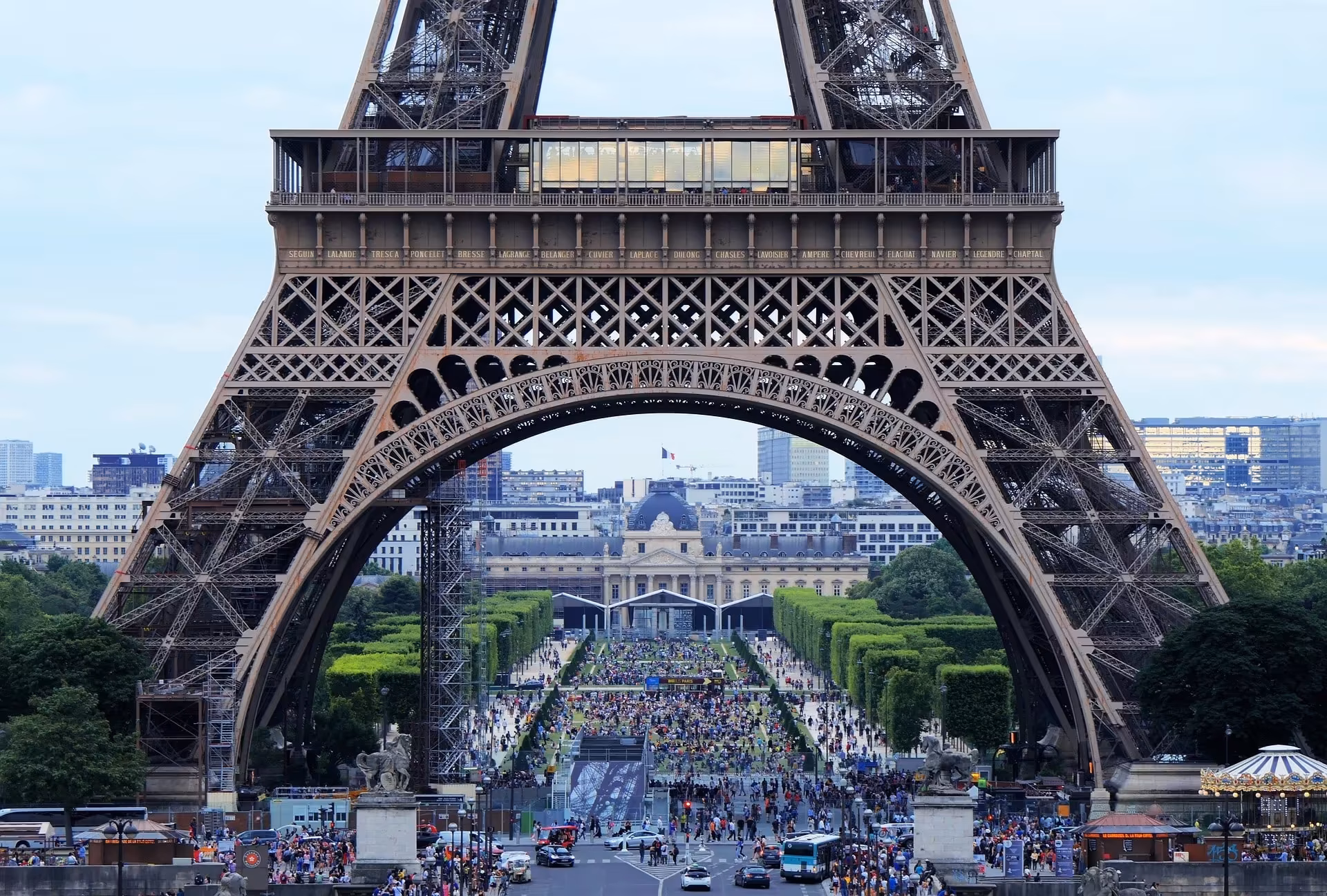 Blick durch den Eiffelturm auf die Parkanlage Champ de Mars in Paris mit zahlreichen Besucherinnen und Besuchern.
