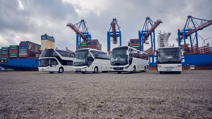 Vier weiße Busse stehen vor großen Hafenkränen und gestapelten Containern.