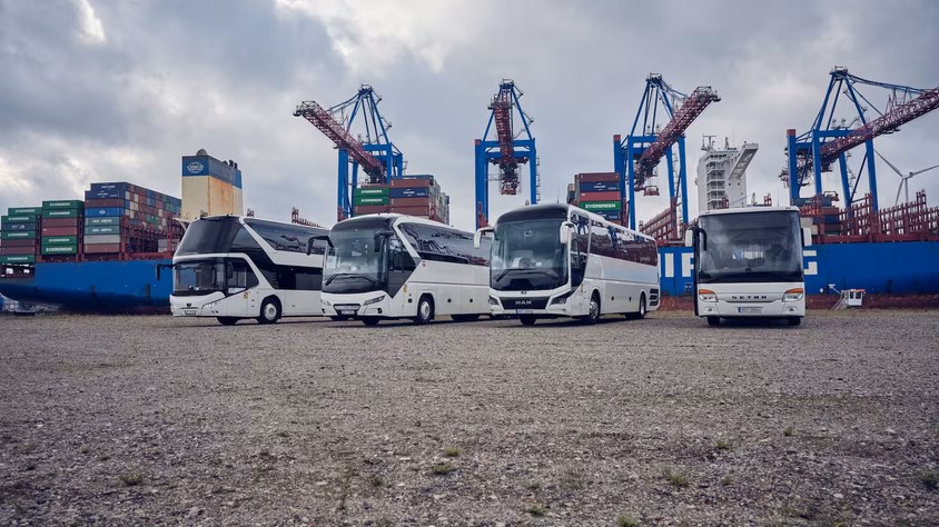Vier weiße Busse stehen vor großen Hafenkränen und gestapelten Containern.
