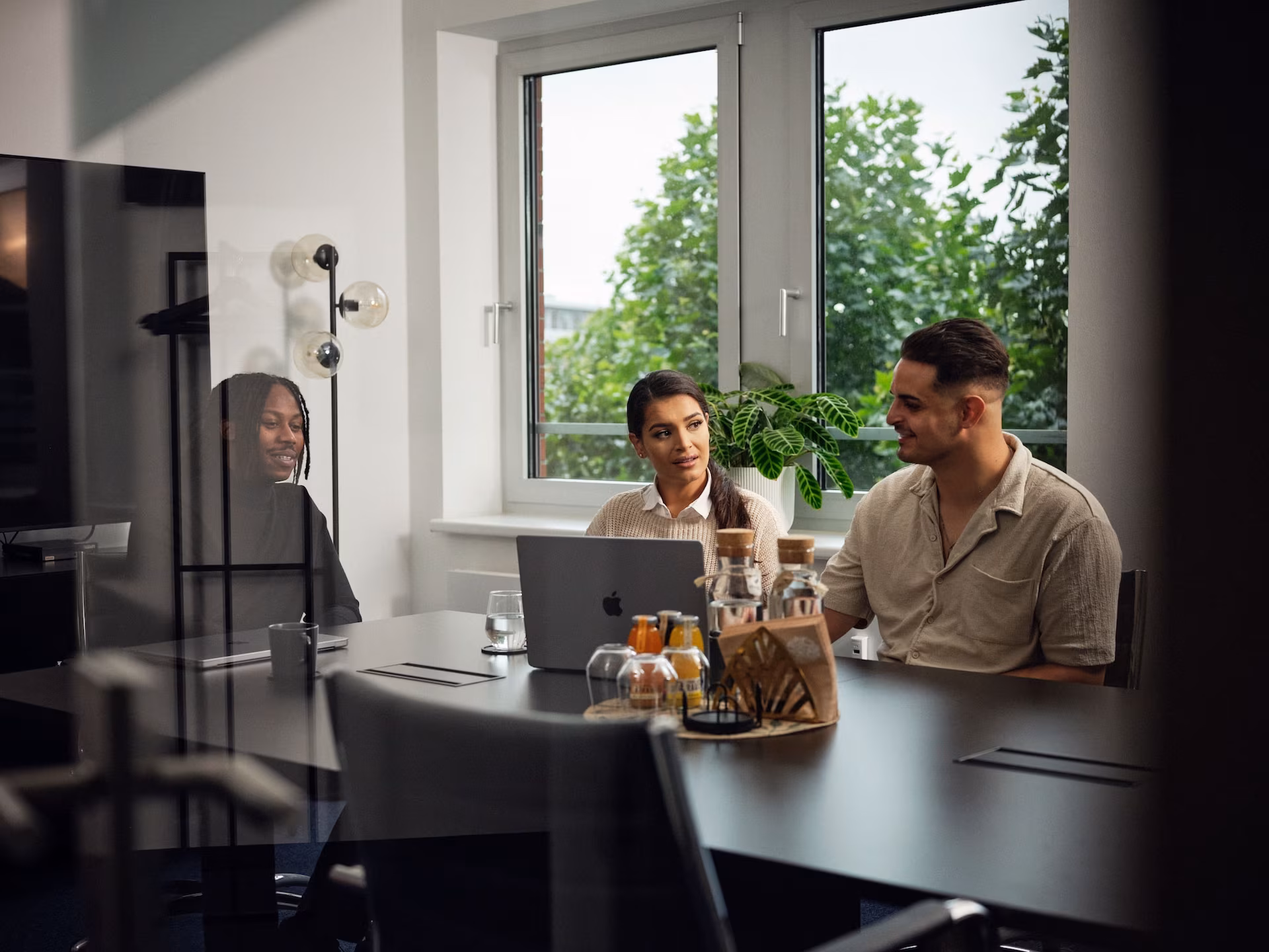 Drei Personen sitzen gemeinsam an einem Besprechungstisch und unterhalten sich vor einem Laptop in einem modernen, hellen Büro mit großen Fenstern und Pflanzen.