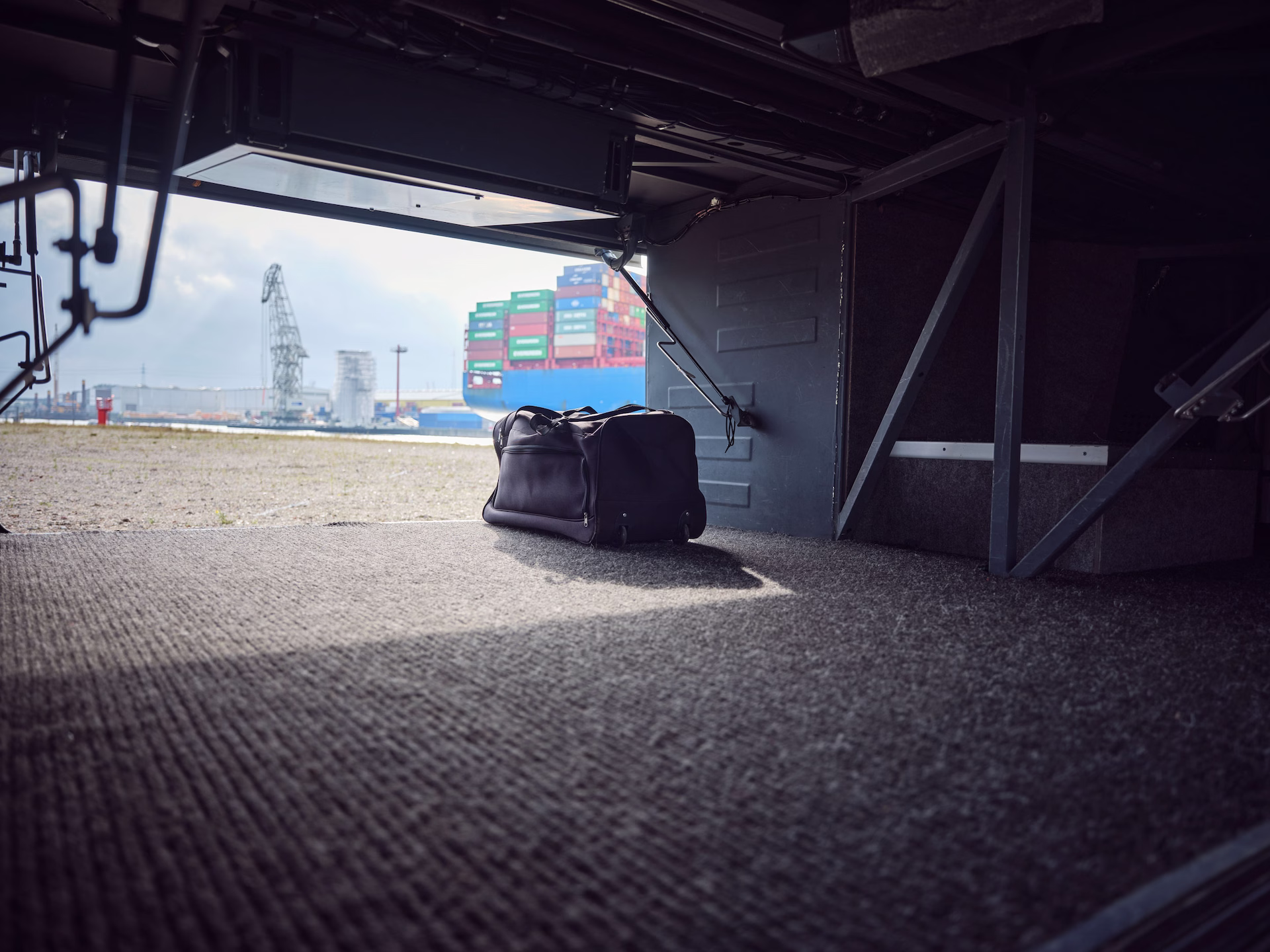 Blick aus dem geöffneten Kofferraum eines Reisebusses, in dem eine dunkle Reisetasche steht; im Hintergrund ist der Hamburger Hafen mit einem Containerschiff und einem Hafenkran zu sehen.