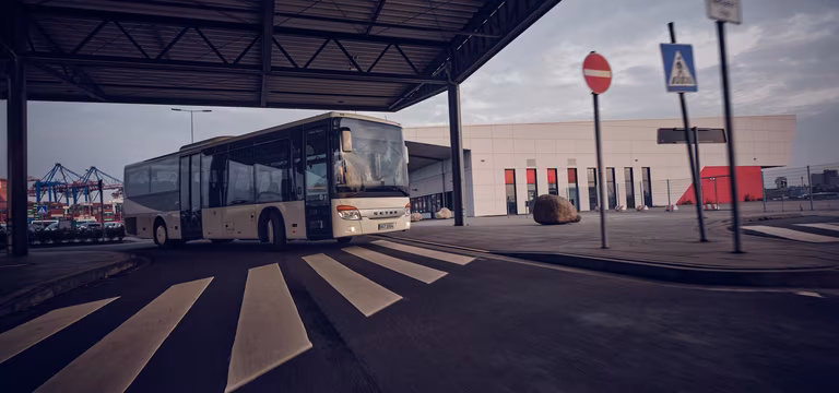 Linienbus fährt unter einer überdachten Zufahrt über einen Zebrastreifen vor einem modernen Gebäudekomplex.