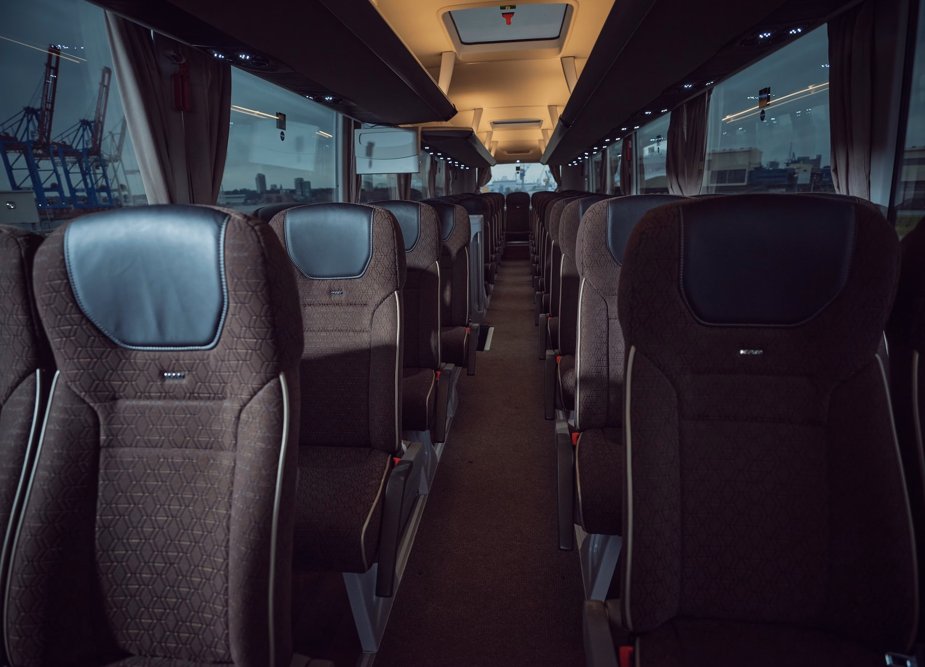 Innenraum eines leeren, modernen Reisebusses mit Reihen dunkler, gepolsterter Sitze und großen Fenstern, durch die eine industrielle Landschaft zu sehen ist.