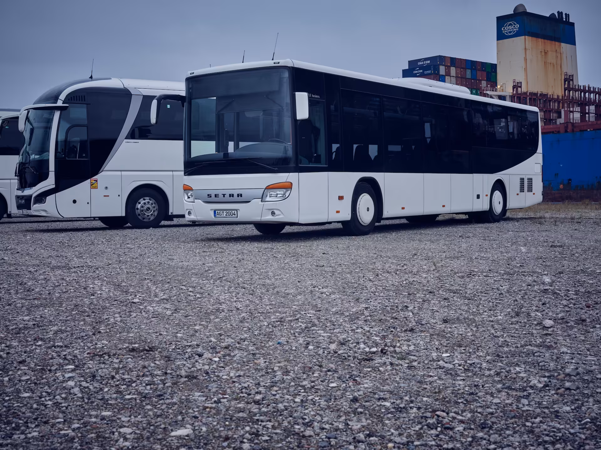 Zwei weiße Busse aus der AGT-Fahrzeugflotte stehen auf einem Schotterplatz, mit Containern im Hintergrund unter einem bewölkten Himmel.