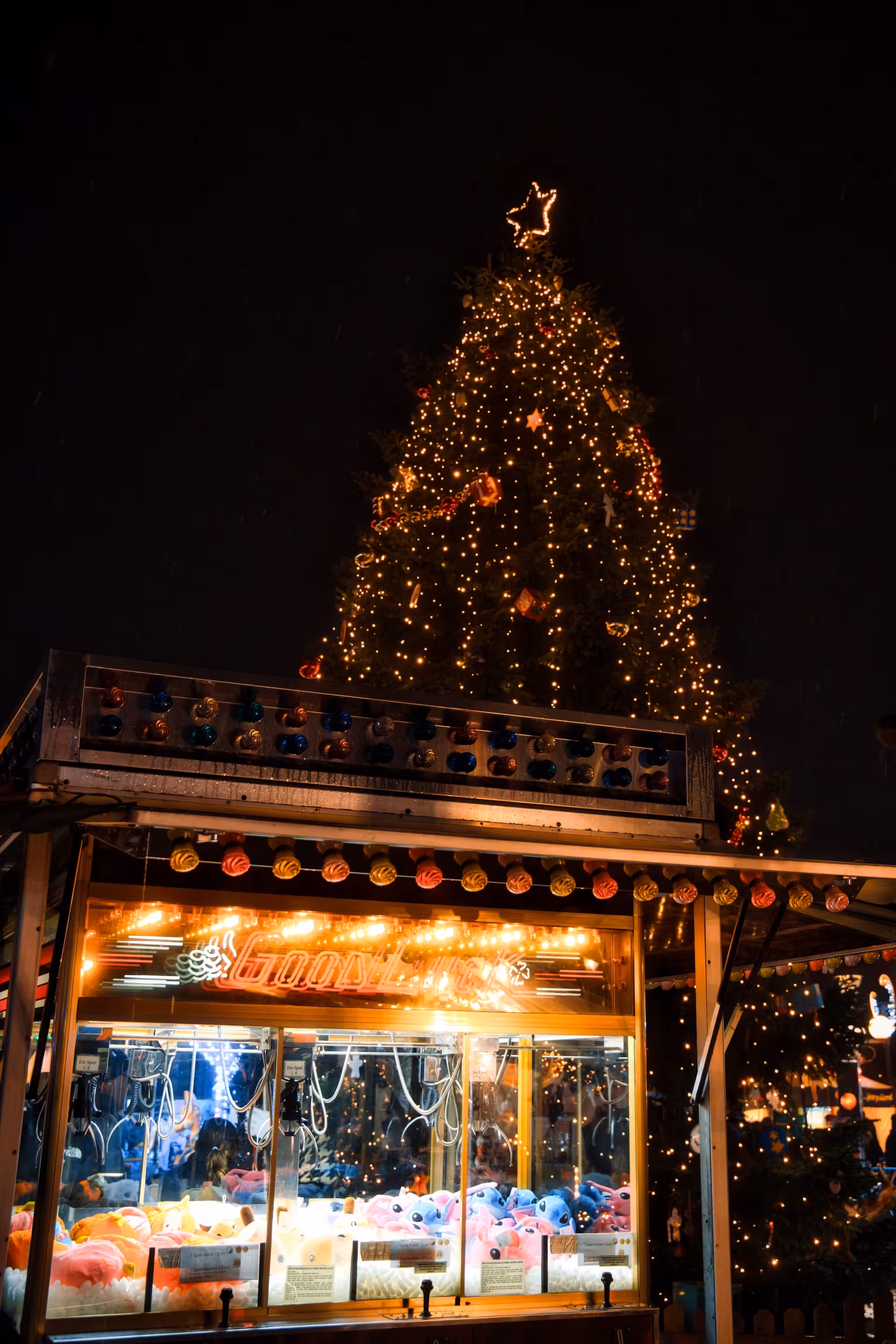 Beleuchteter Greifautomat mit Plüschtieren auf einem nächtlichen Weihnachtsmarkt, dahinter ein großer, mit Lichterketten und Stern geschmückter Weihnachtsbaum.