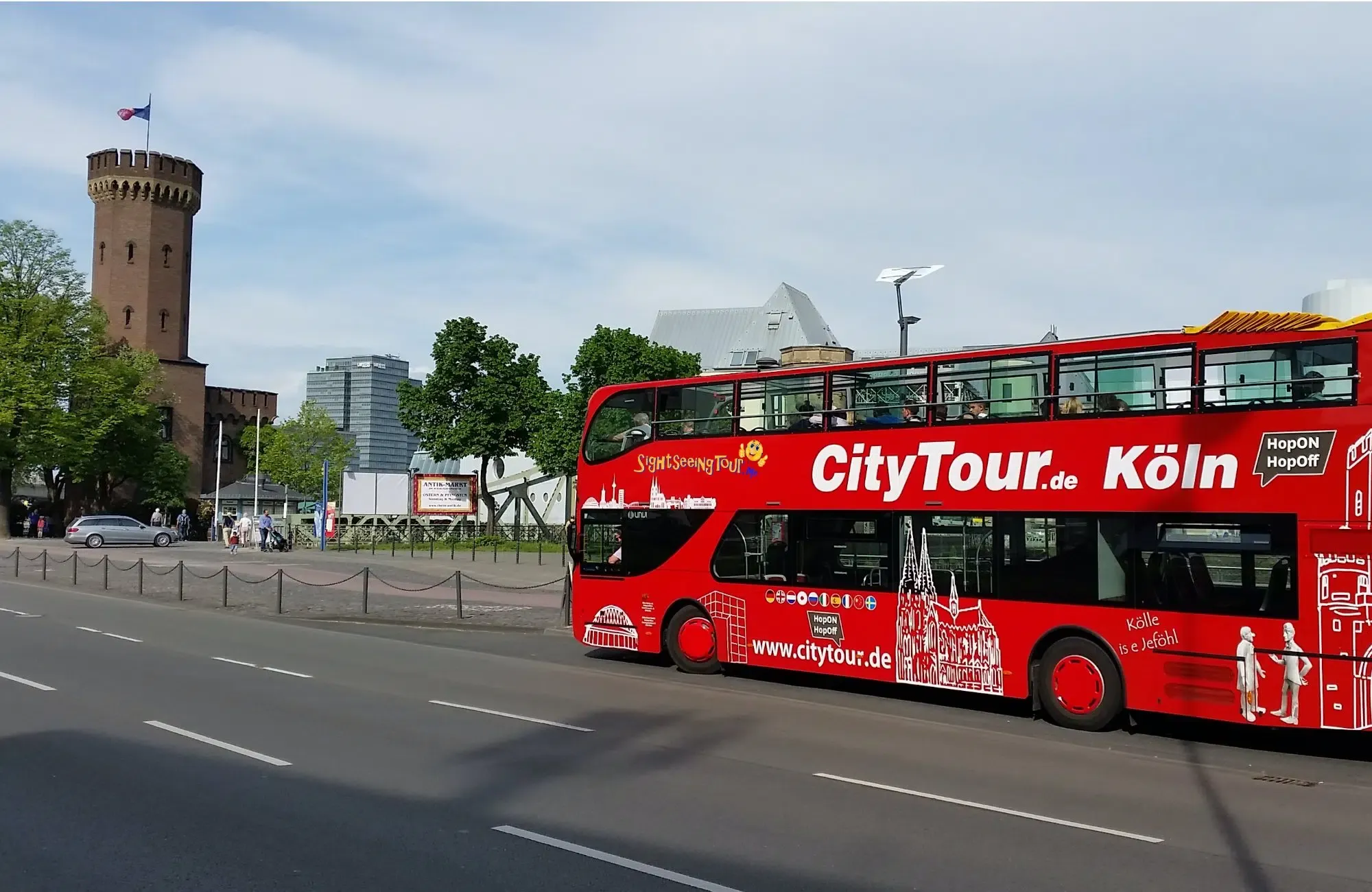 Ein roter „CityTour.de Köln“ Hop-On Hop-Off Doppeldeckerbus fährt auf einer Straße in Köln, im Hintergrund sind der historische Malakoffturm sowie moderne Stadtgebäude zu sehen.