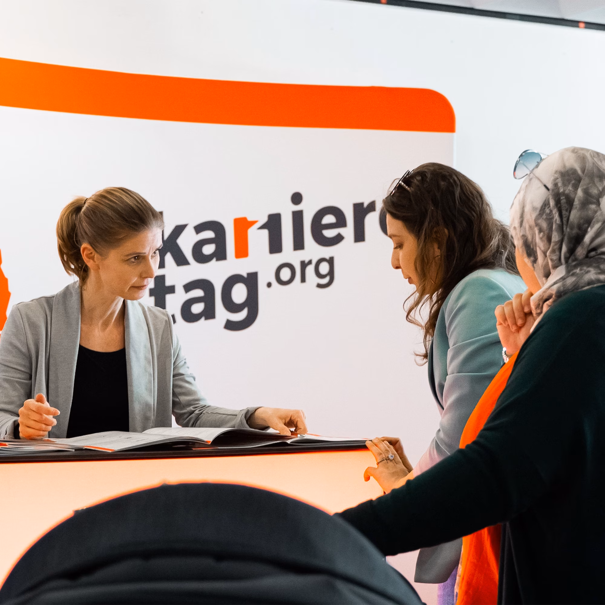 Mitarbeiterin berät zwei Besucherinnen an einem Messestand zum Thema Karriere und Ausbildung, vor einem Informationsbereich mit der Aufschrift „Karrieretag.org“.