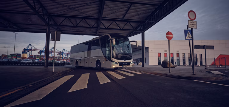 Ein weißer Reisebus fährt unter einer großen Überdachung hervor auf einen Zebrastreifen zu, im Hintergrund sind Hafenkräne und Verkehrszeichen erkennbar.