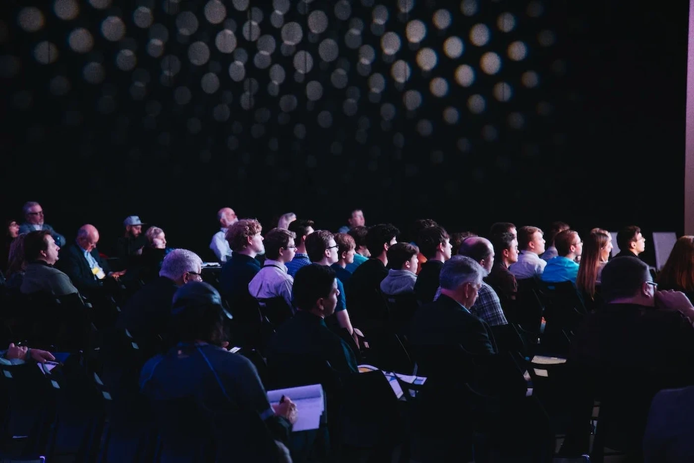 Audience seated in a dimly lit room, attentively facing a stage. Circular lights create a pattern on the dark wall behind them.
