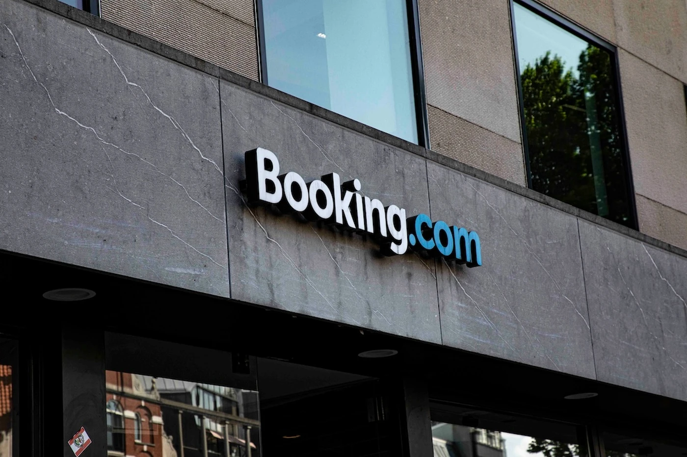 Building facade with a "Booking.com" sign in white and blue letters, reflecting urban surroundings in the windows above.