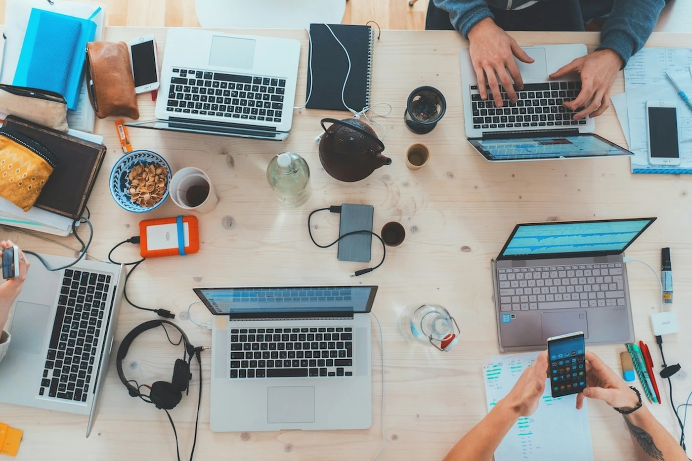 Aerial view of a cluttered desk with laptops, phones, notebooks, headphones, and snacks, indicating a busy workspace with multiple people.