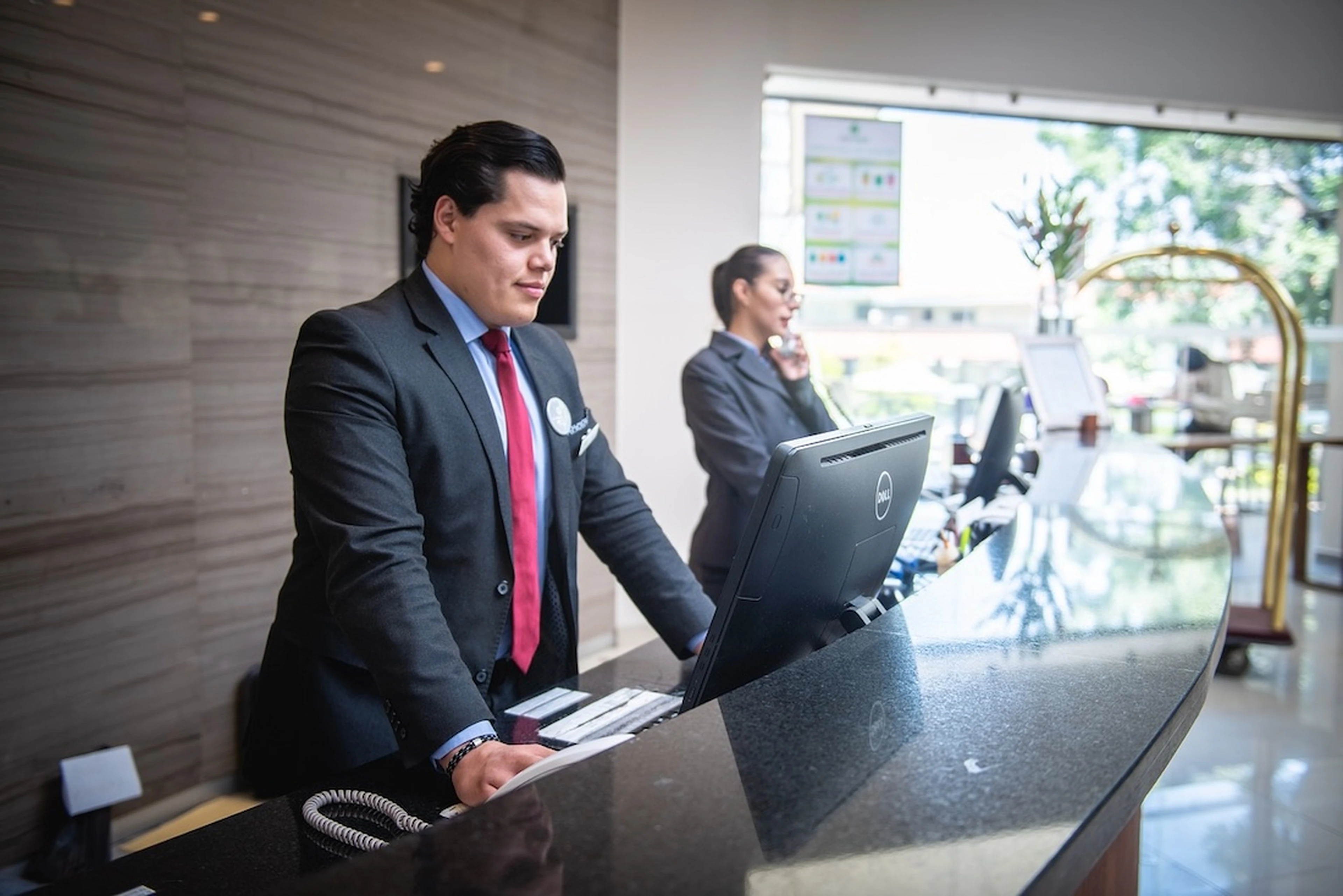 Hotel receptionists working at a modern front desk, one using a computer and the other on the phone, with a bright lobby in the background, highlighting exemplary customer service