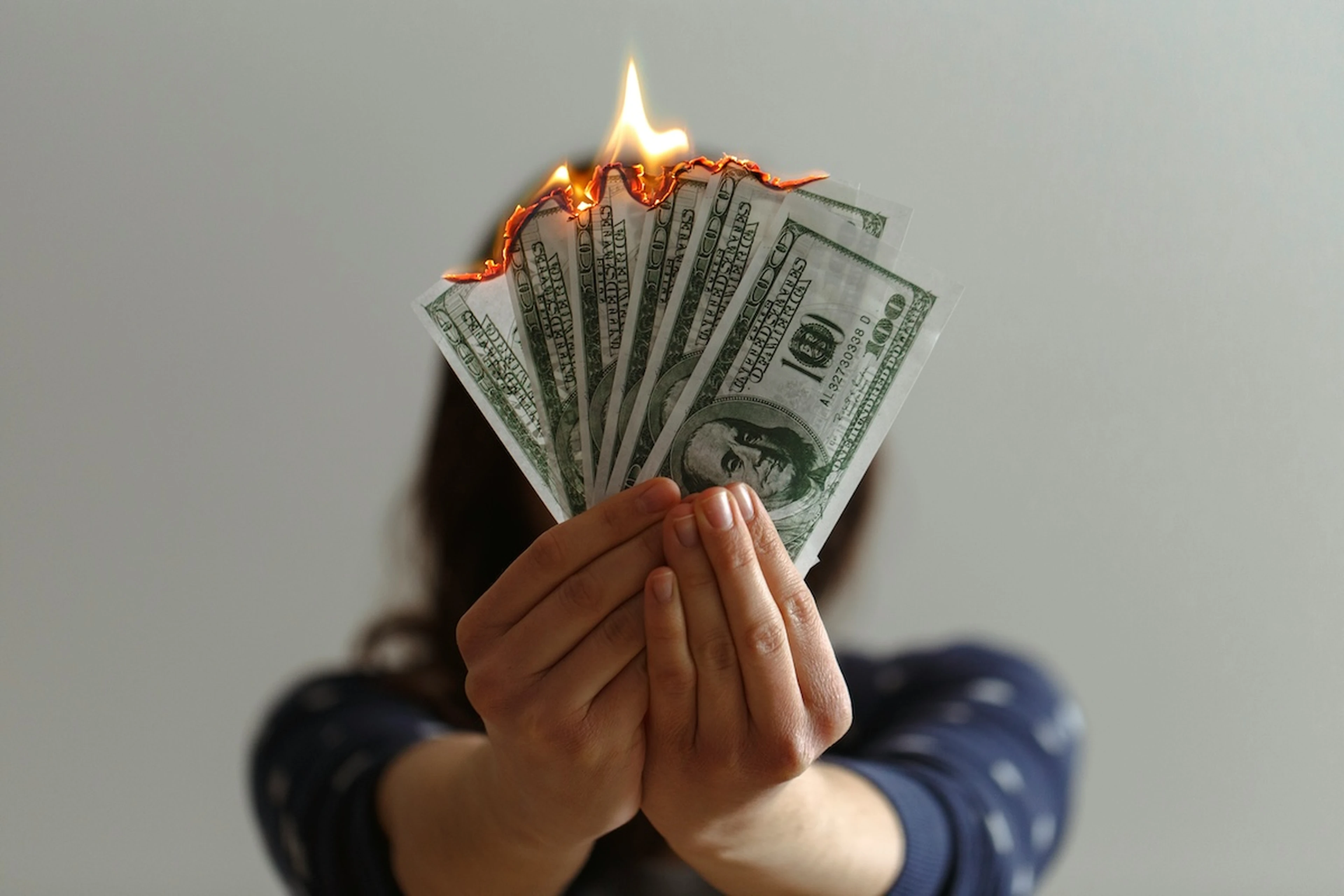 Person holds a fanned stack of burning $100 bills, flames licking along the top edge.