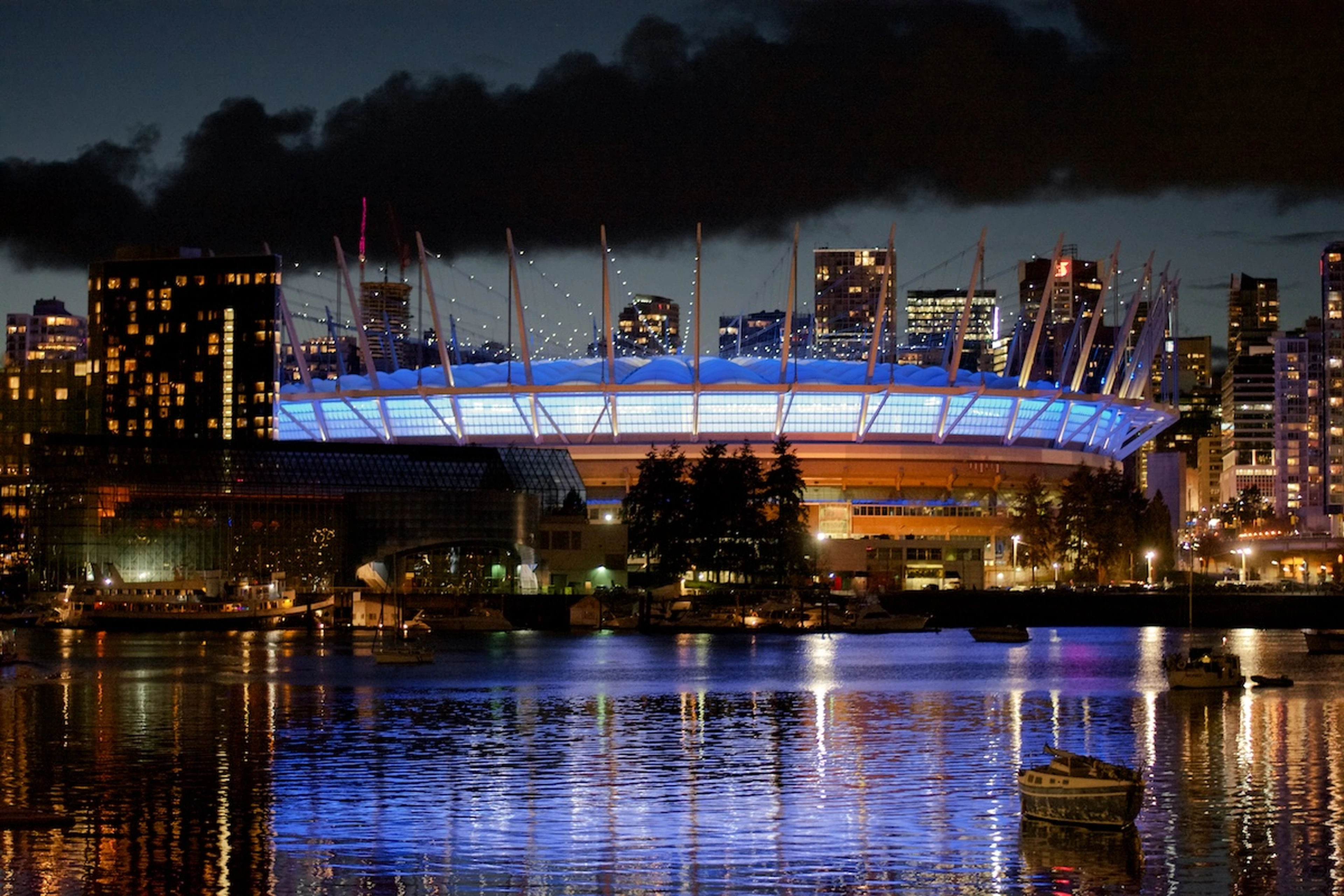 Blue-lit waterfront stadium In vancouver with spire-like masts at night, city skyline and clouds behind, reflections shimmering on the water, boats in foreground.
