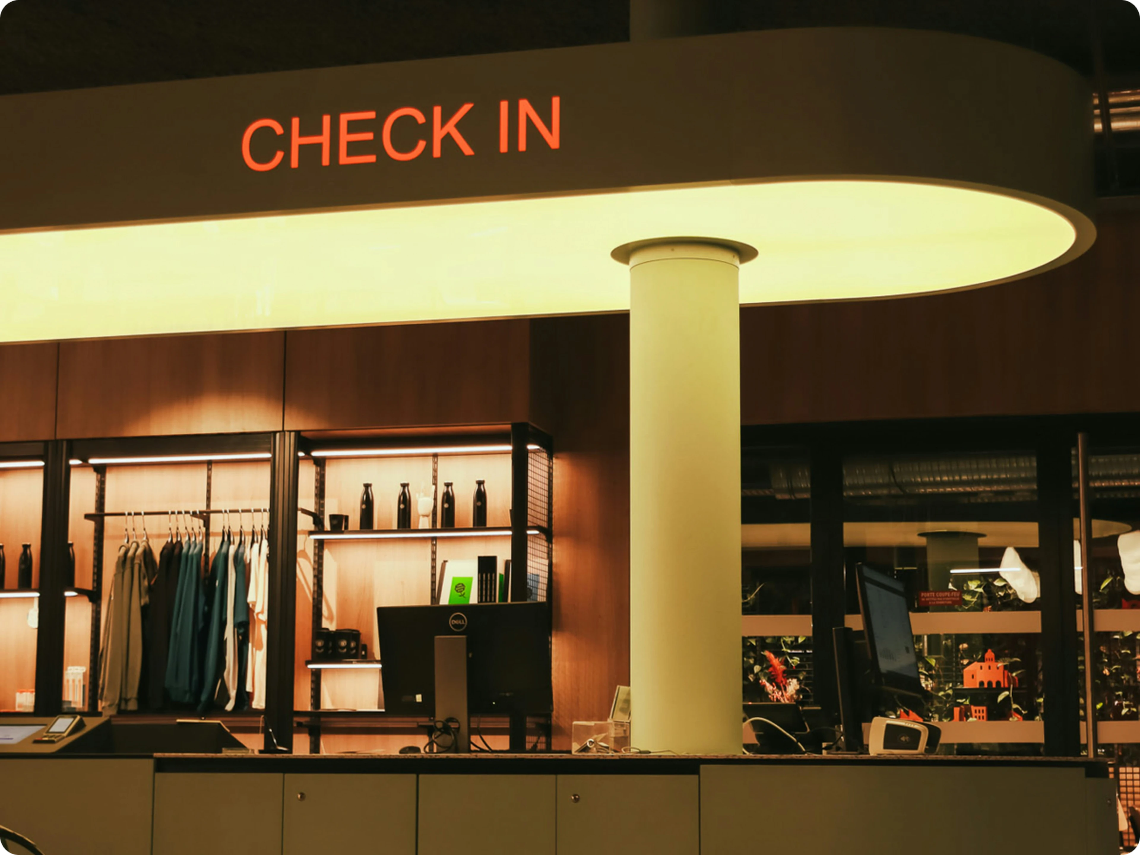 Hotel check-in counter with illuminated sign, computers, and shelves displaying clothing and bottles. Warm lighting creates a welcoming atmosphere.
