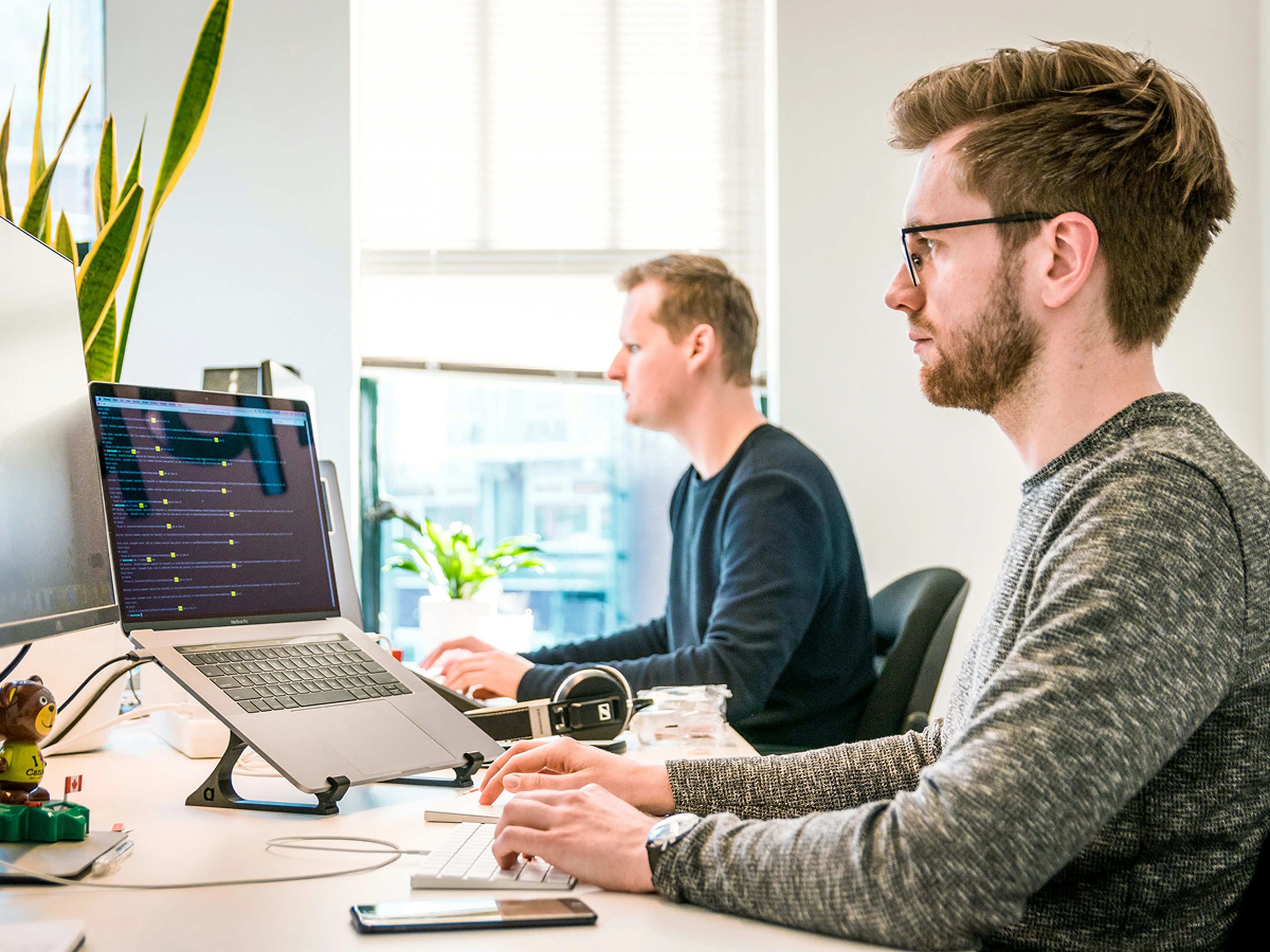 Two men working at desks with laptops in a bright office, one focused on his screen with rate insights, the other typing, with a plant nearby.