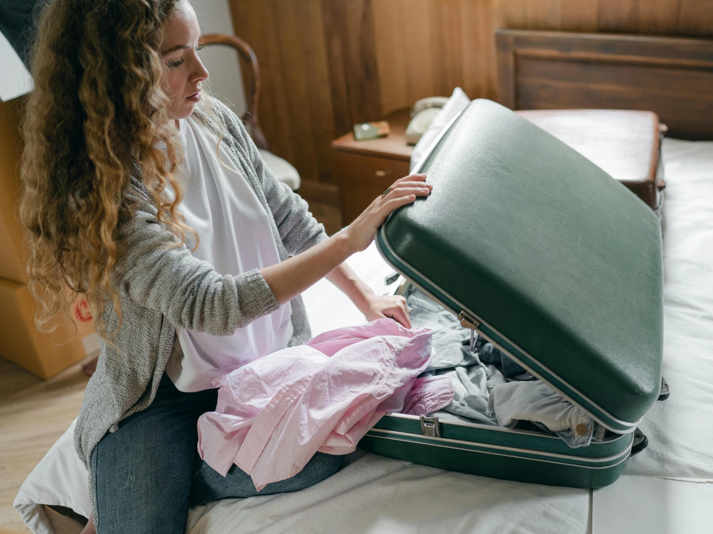 Woman sitting on a bed packing clothes into an open green suitcase.