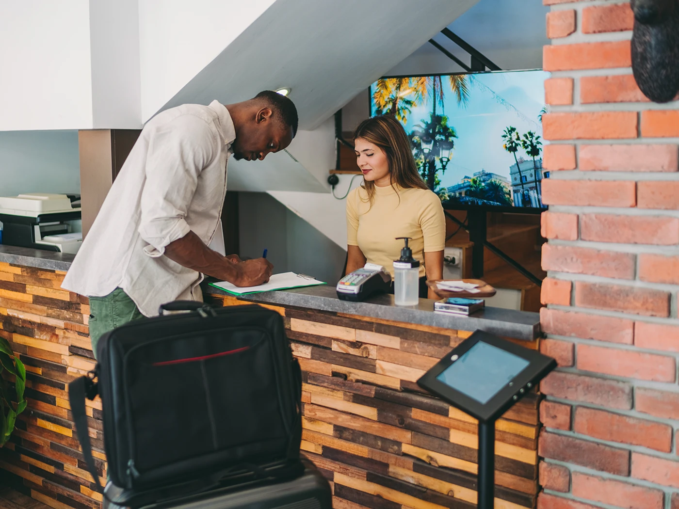 Man signing in at a hotel reception, with a suitcase beside him. A woman stands behind the desk, and a TV displays a tropical scene.
