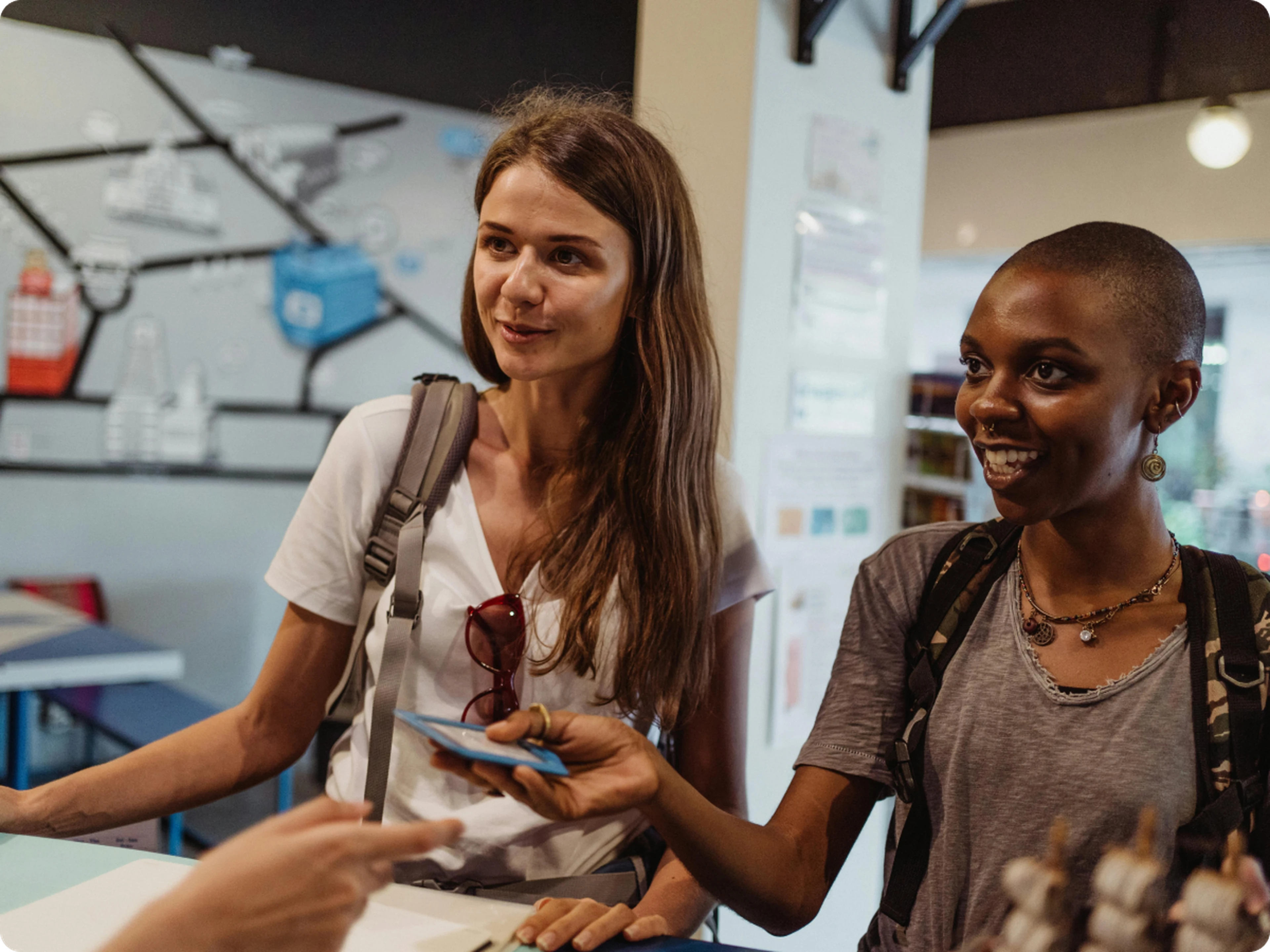 Two young women with backpacks smile at a hotel front desk as one hands a card to a staff member.