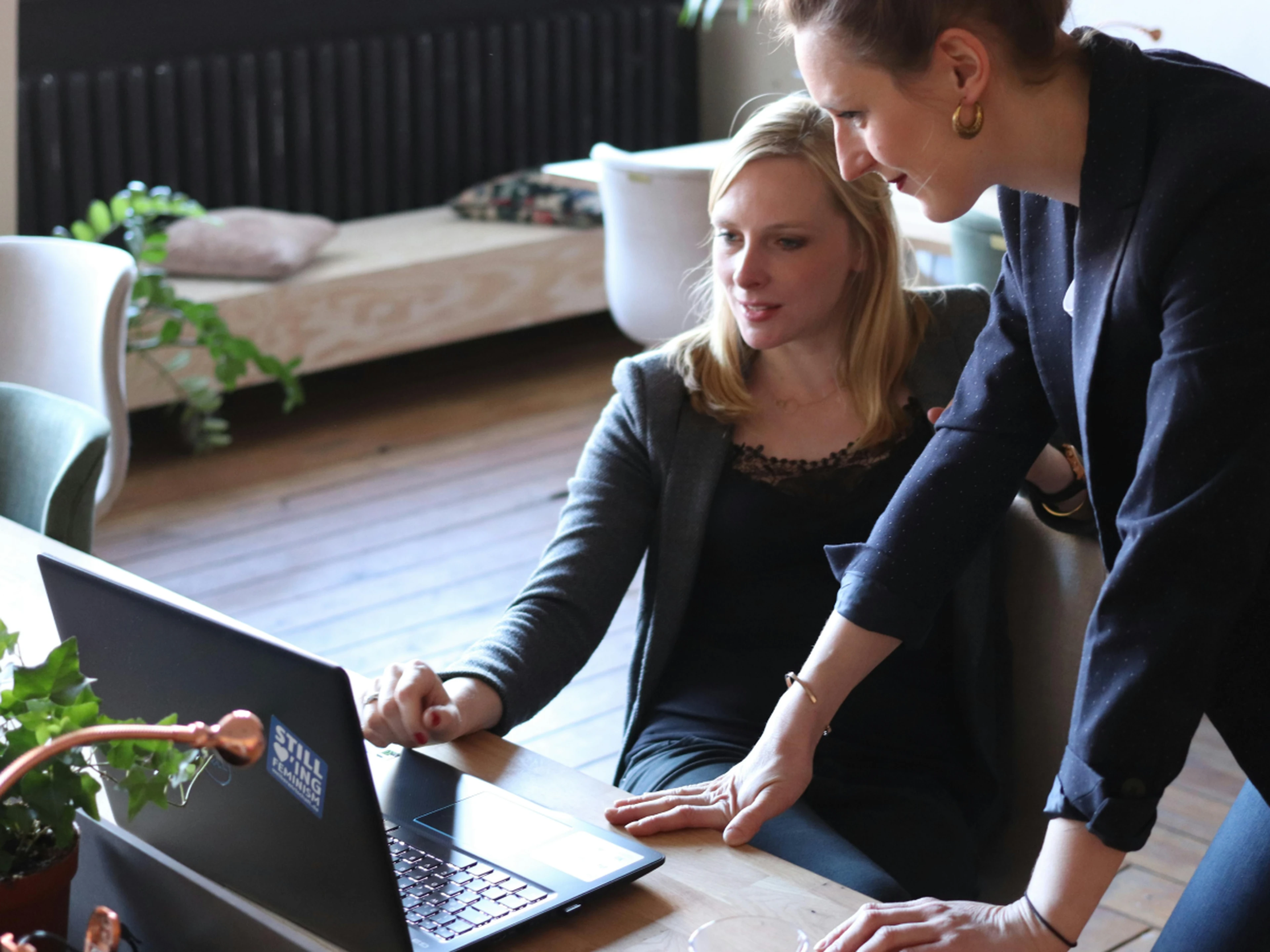 Two colleagues look at a laptop together at a table in a bright office with plants in the background