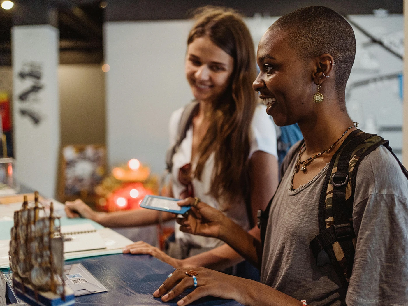 Two women smiling at a counter, one holding a phone. A small ship model is on the counter. Both carry backpacks, suggesting travel or exploration.