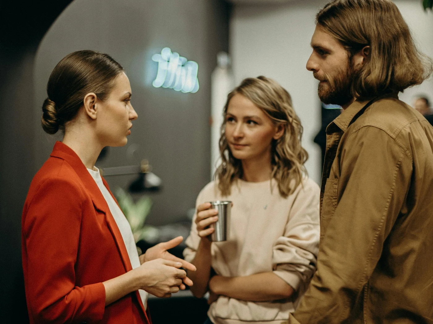 Three people in conversation indoors: a woman in a red blazer speaking to a woman holding a metal cup and a long-haired man listening.