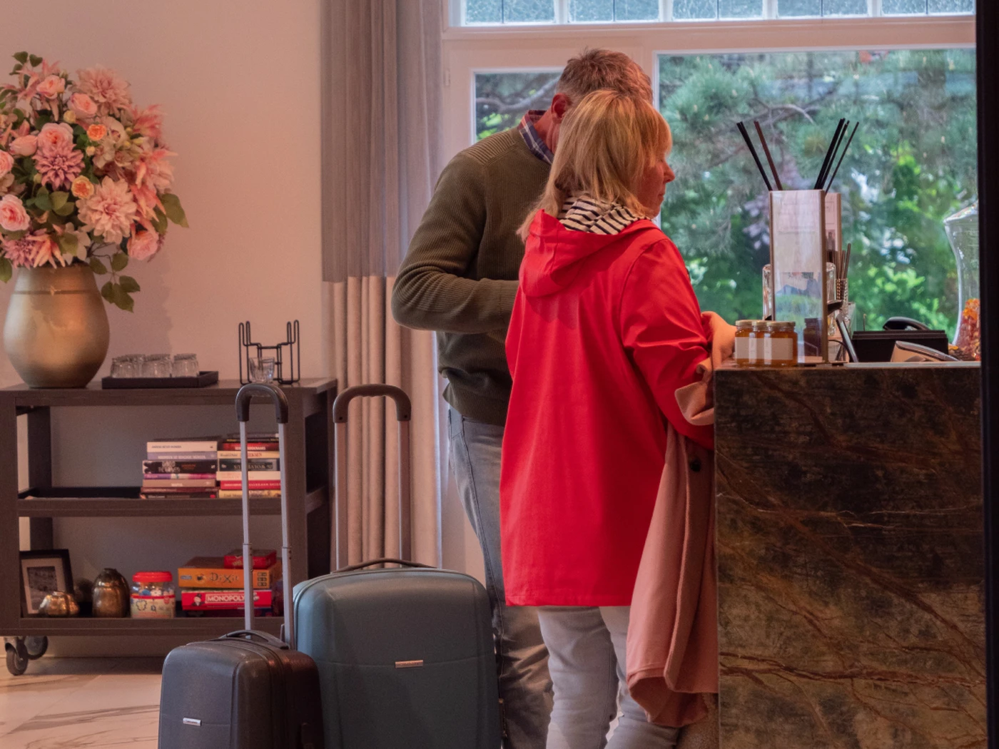 Two travelers with suitcases at a hotel reception desk; woman in red jacket checking in, man beside her and floral arrangement on a shelf.