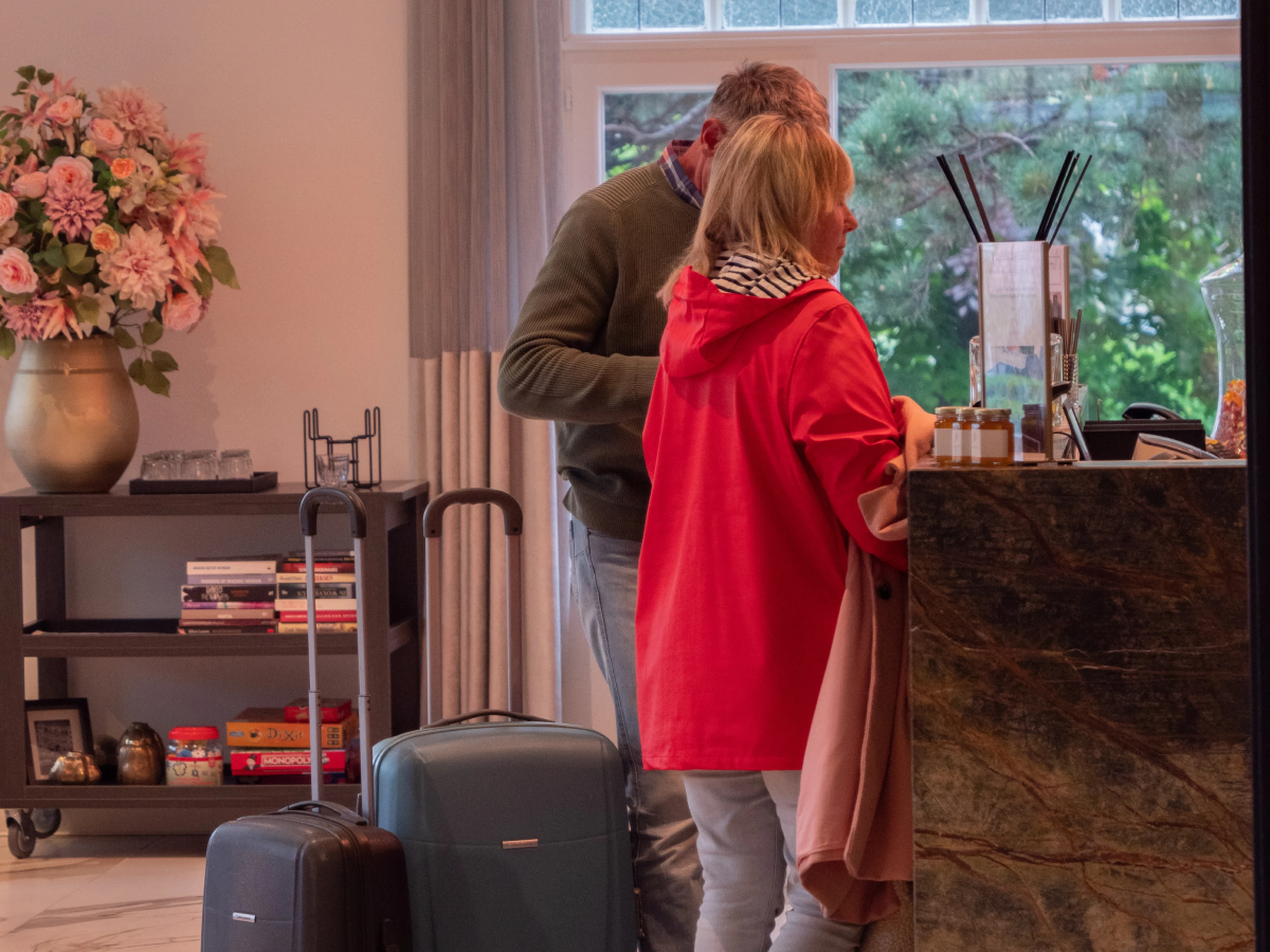 Two travelers with suitcases at a hotel reception desk; woman in red jacket checking in, man beside her and floral arrangement on a shelf.