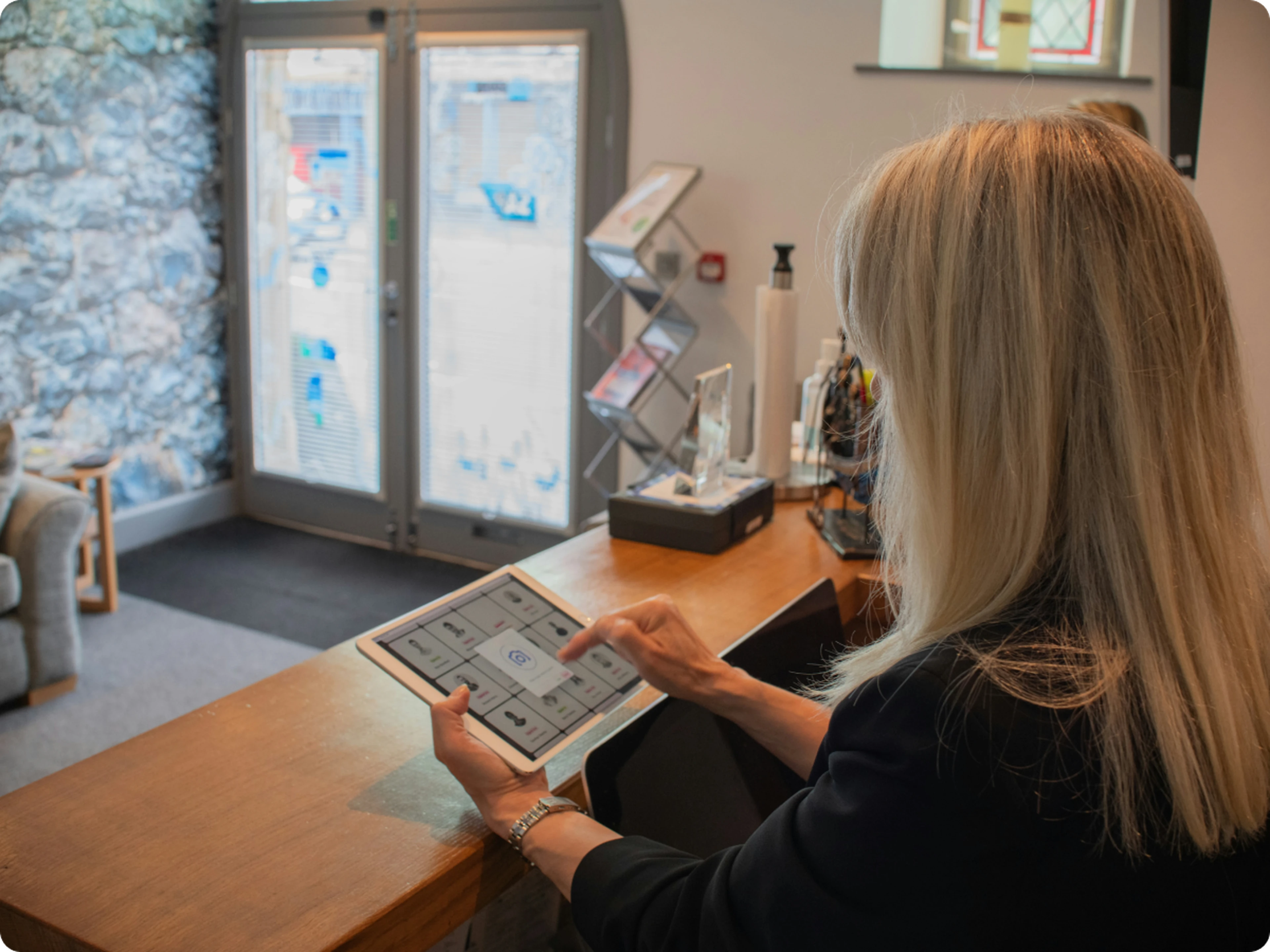 Woman with long blonde hair using a tablet at a hotel reception desk by glass doors.