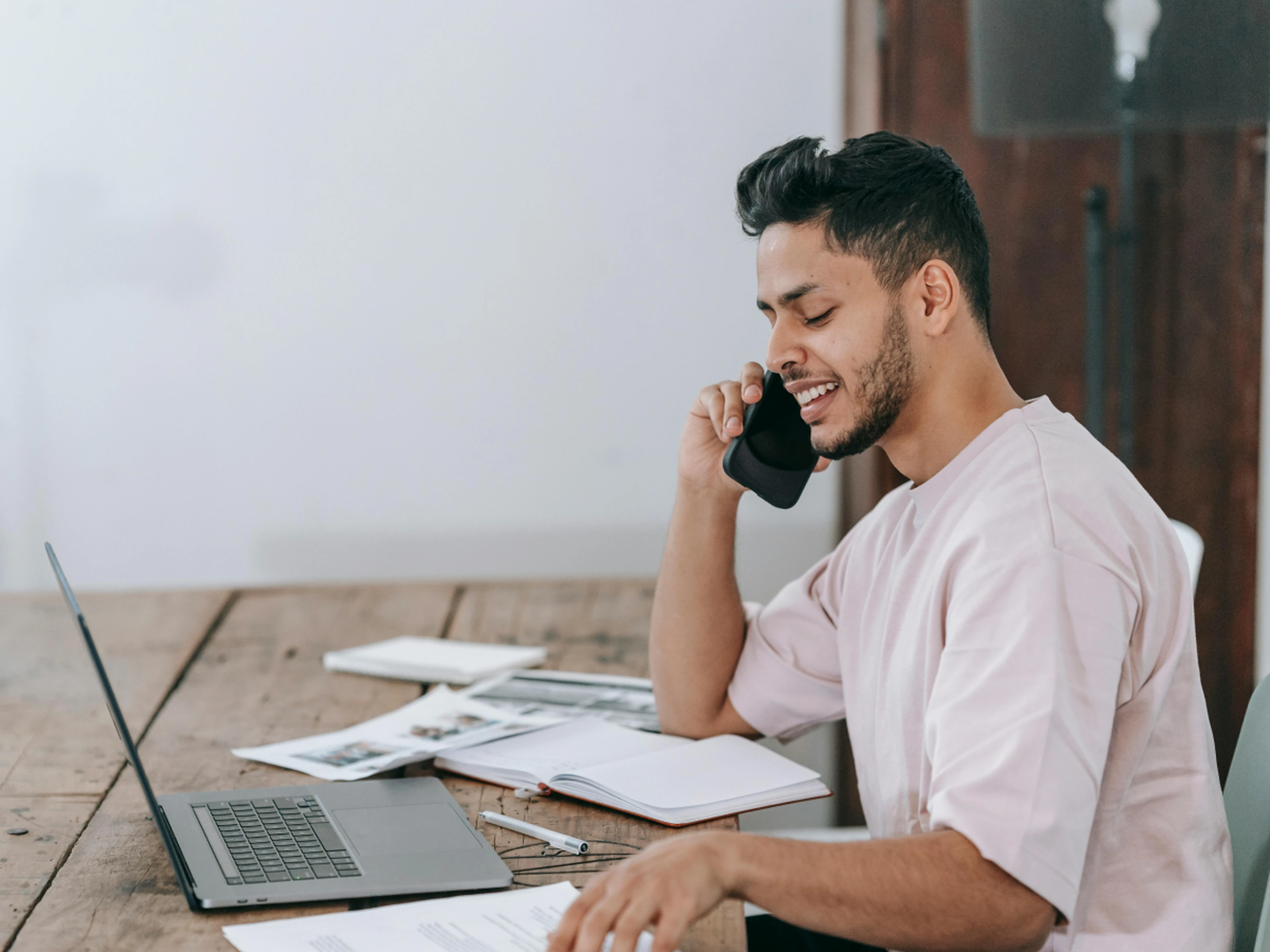 Happy man calling whilst looking at laptop with papers around