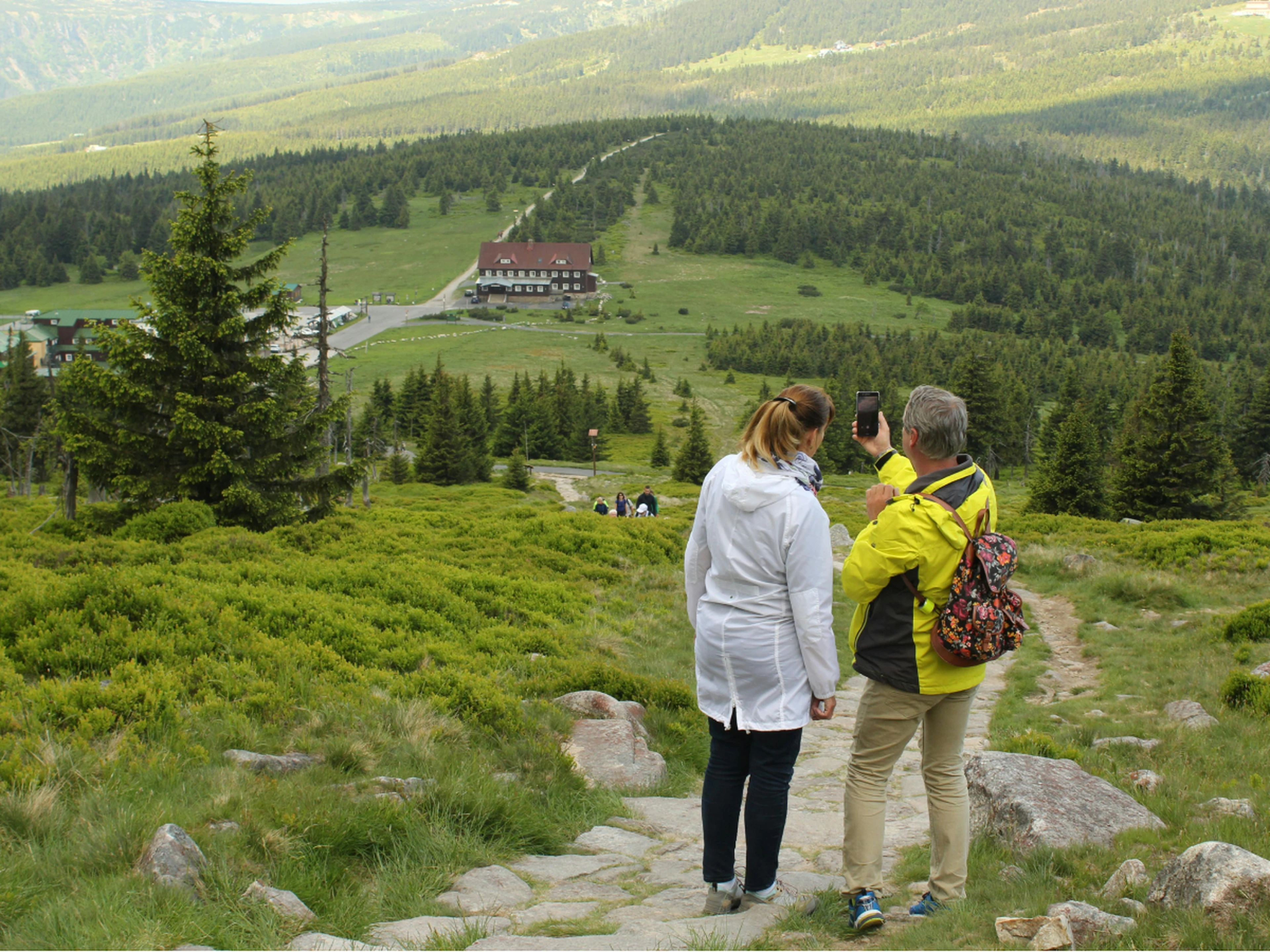 Two local travelers hiking, overlooking a green valley with trees and a mountain lodge