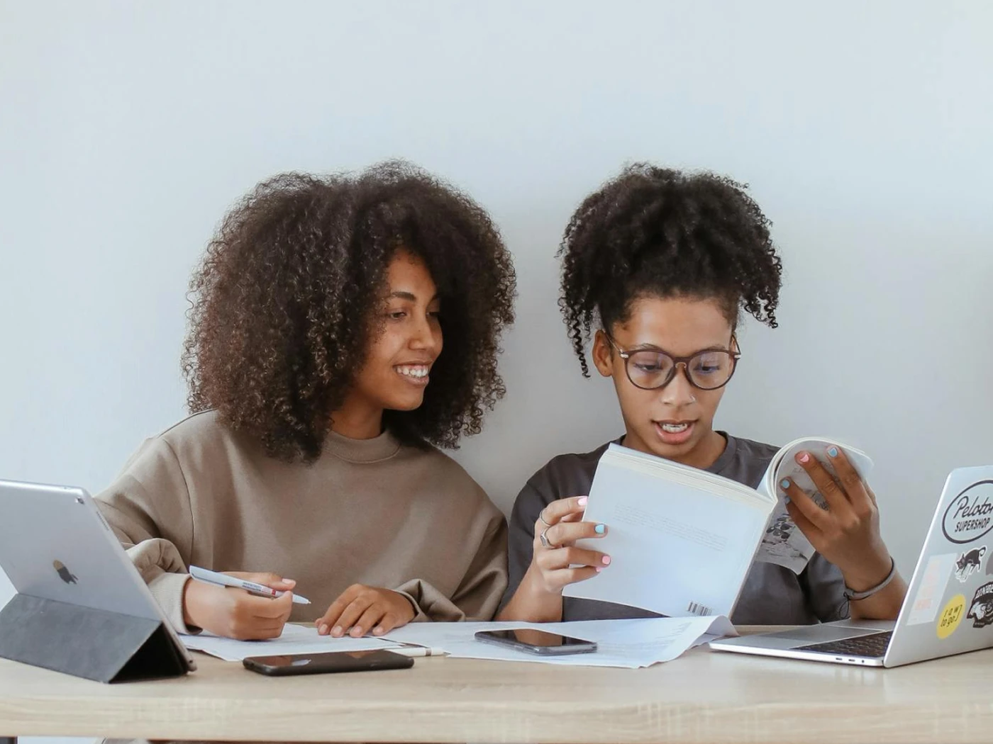 Two women reading a book with a laptop and a tablet in front of them