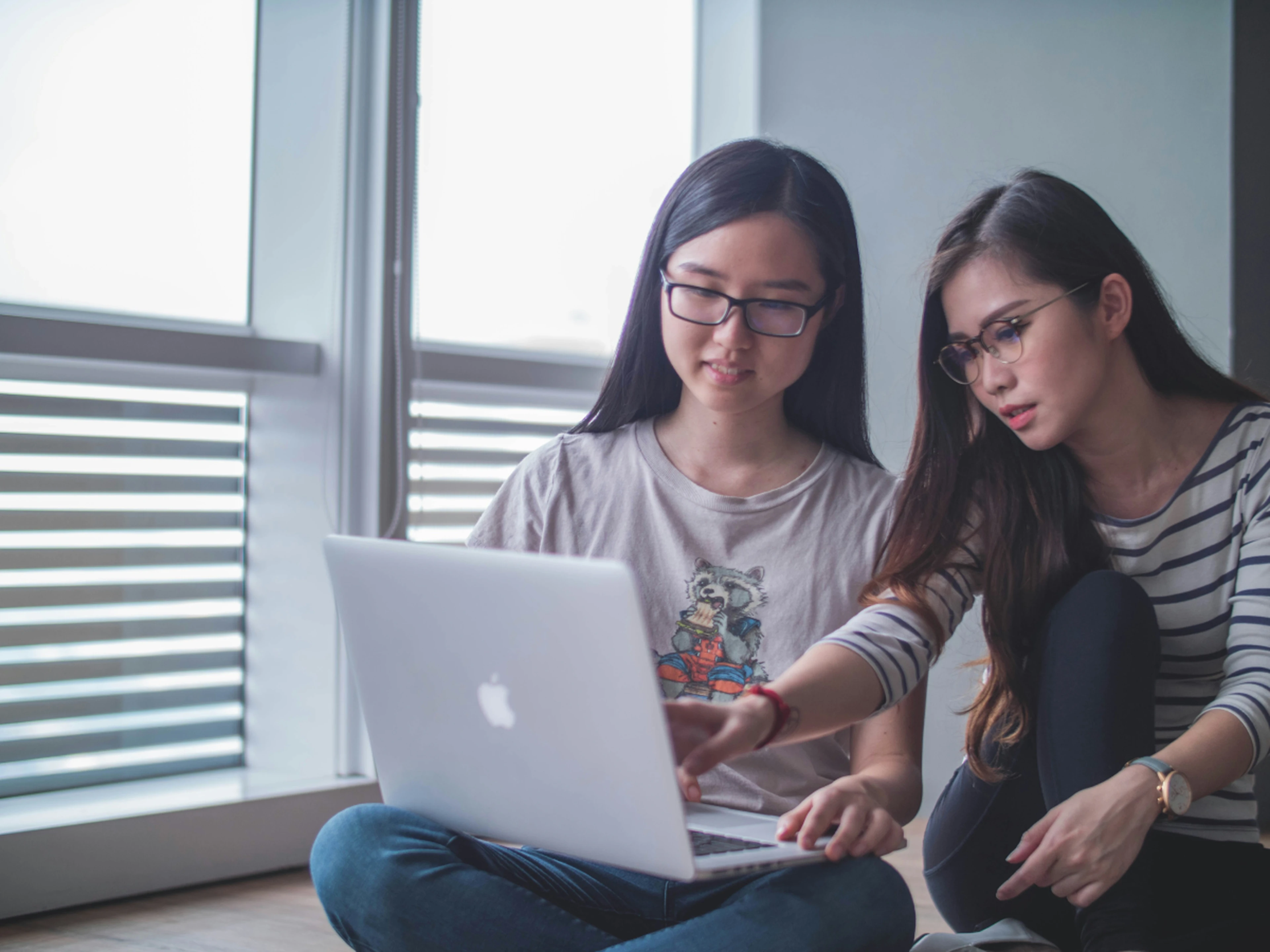 Two women sit on the floor by a window and look at a laptop together, one pointing at the screen