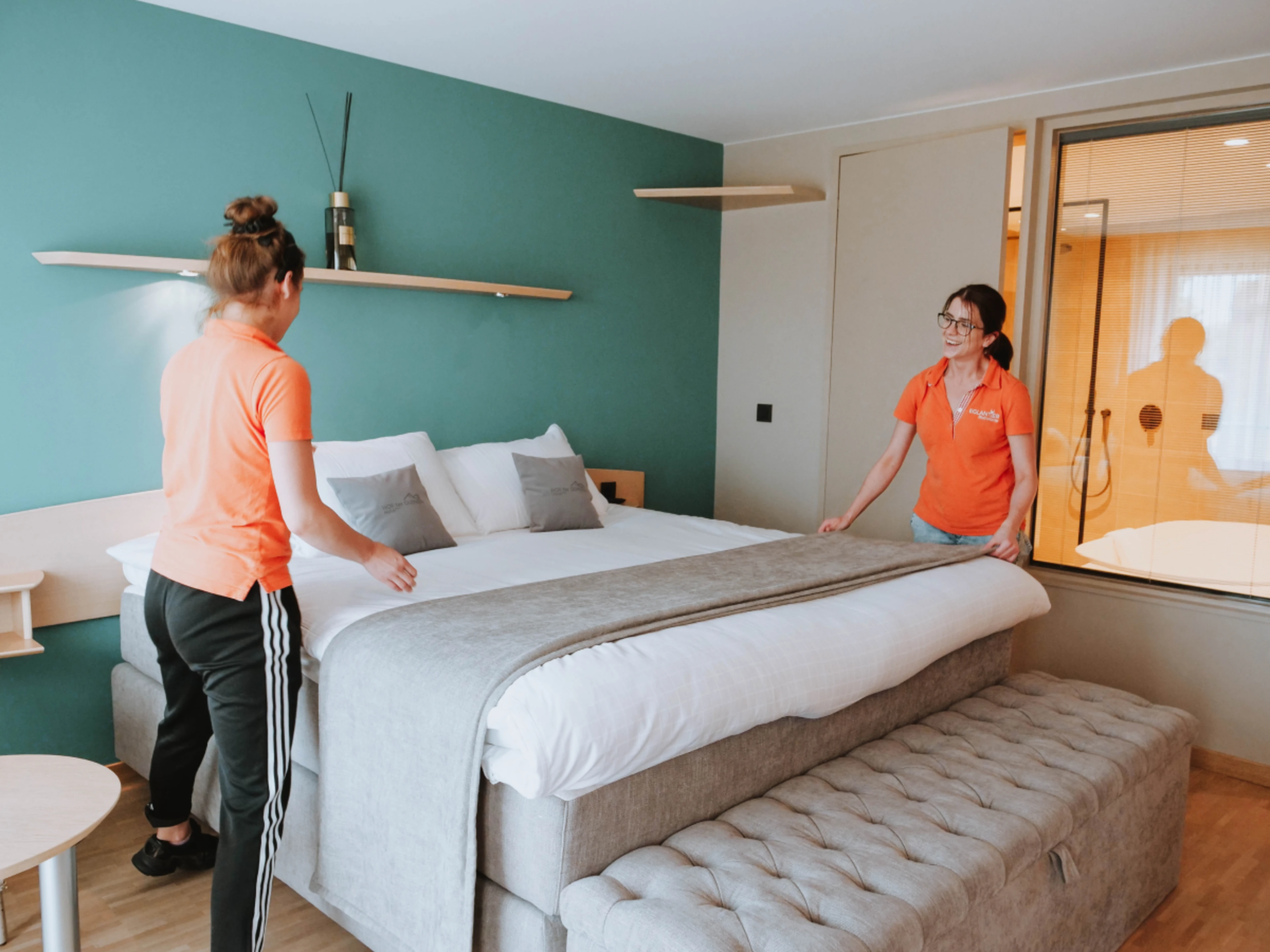 Two housekeepers in orange shirts making a bed in a modern hotel room with a teal accent wall and tufted bench