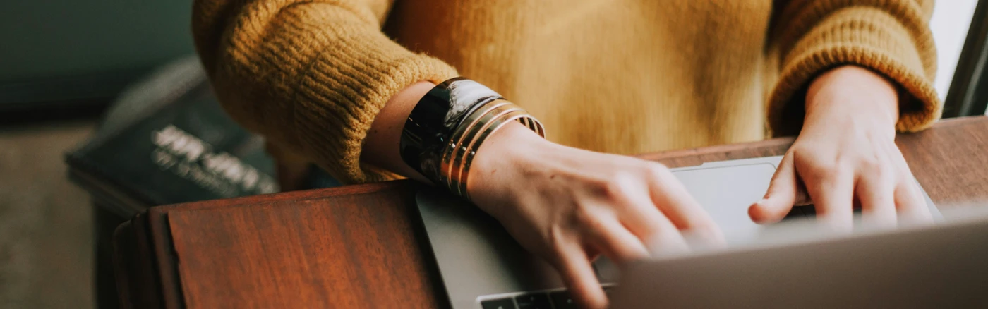 Close-up of hands typing on a laptop at a wooden desk, person wearing a mustard sweater and stacked bracelets.