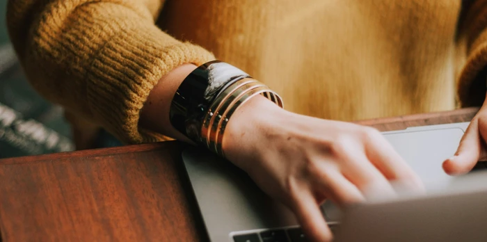 Close-up of hands typing on a laptop at a wooden desk, person wearing a mustard sweater and stacked bracelets.