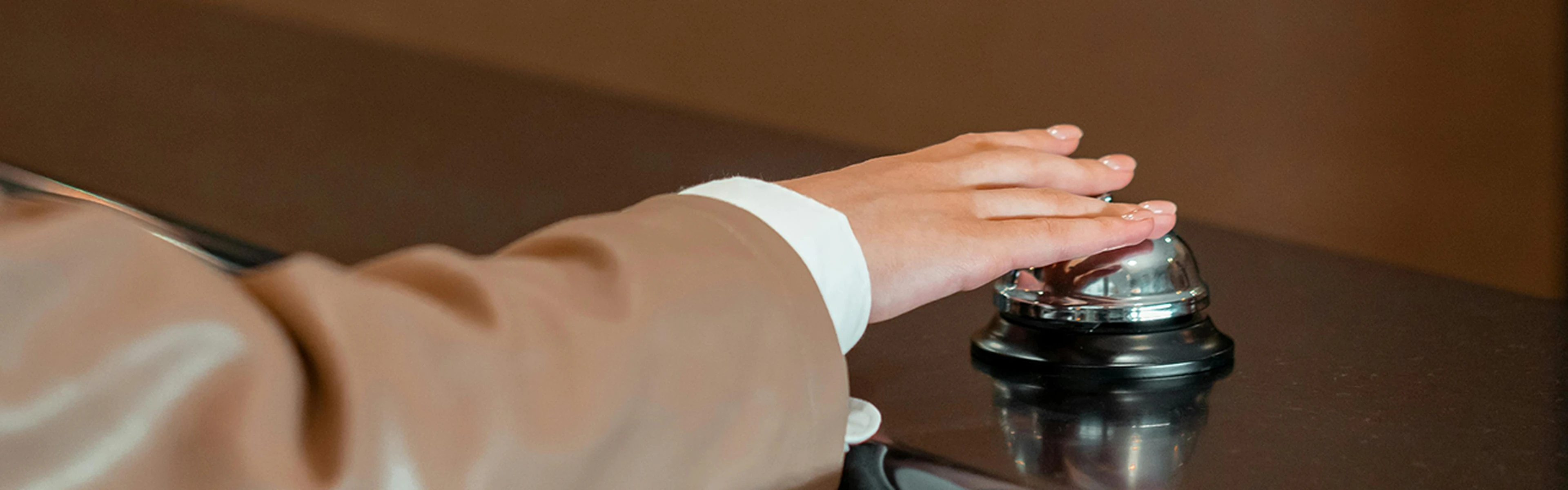A person in a brown suit ringing a silver service bell on a dark countertop.