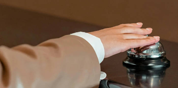 A person in a brown suit ringing a silver service bell on a dark countertop.