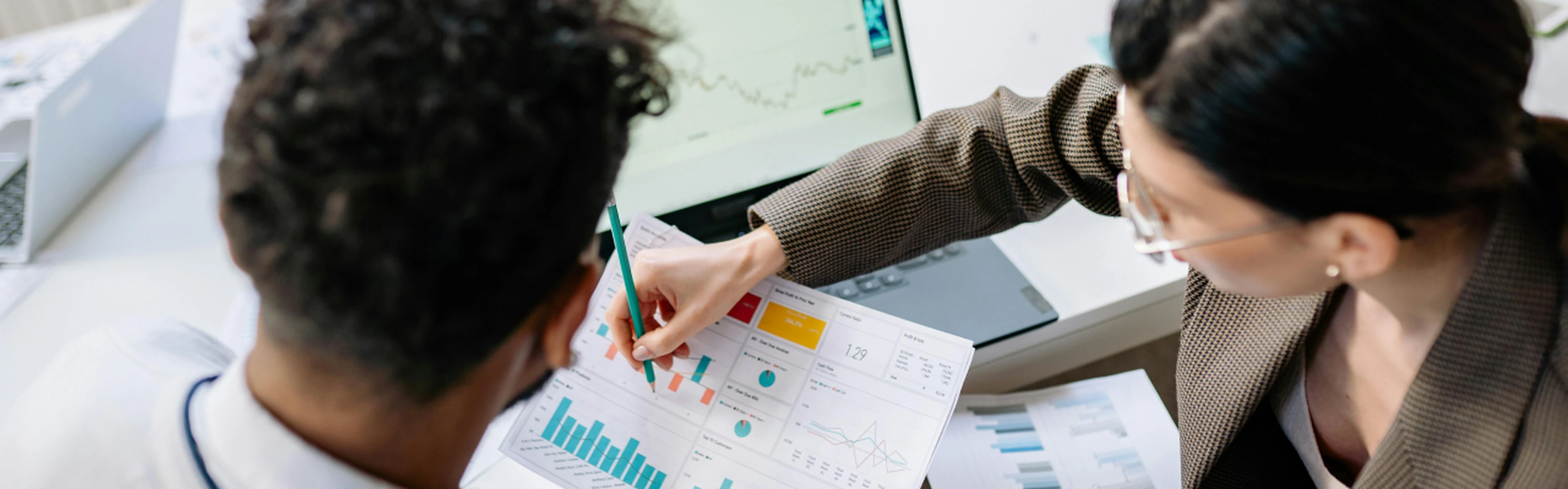 Two professionals review charts and graphs on paper and a laptop, discussing data trends